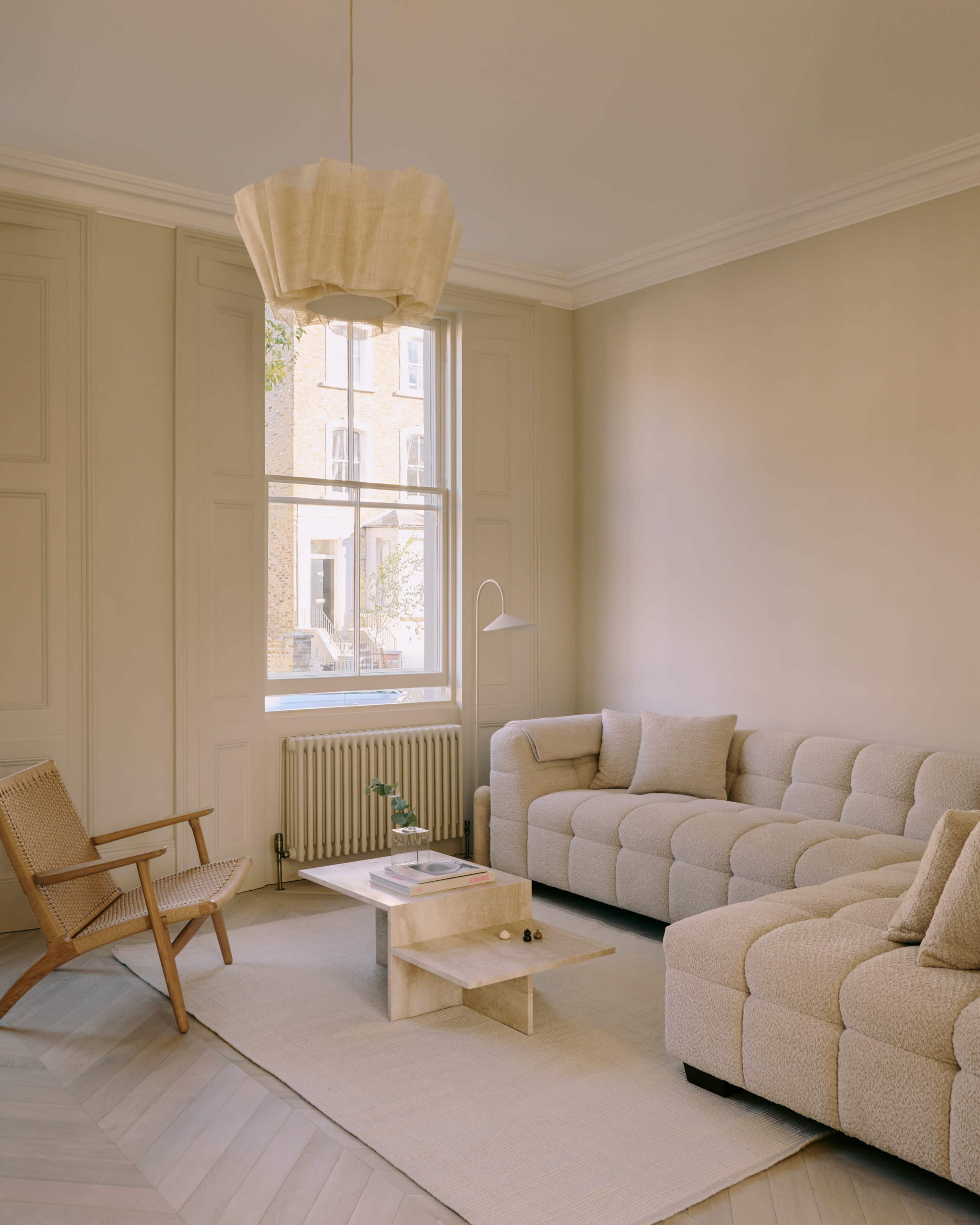 The image features a light-colored living room with a beige sectional sofa, a wooden armchair, a coffee table, and a large window allowing natural light to enter.