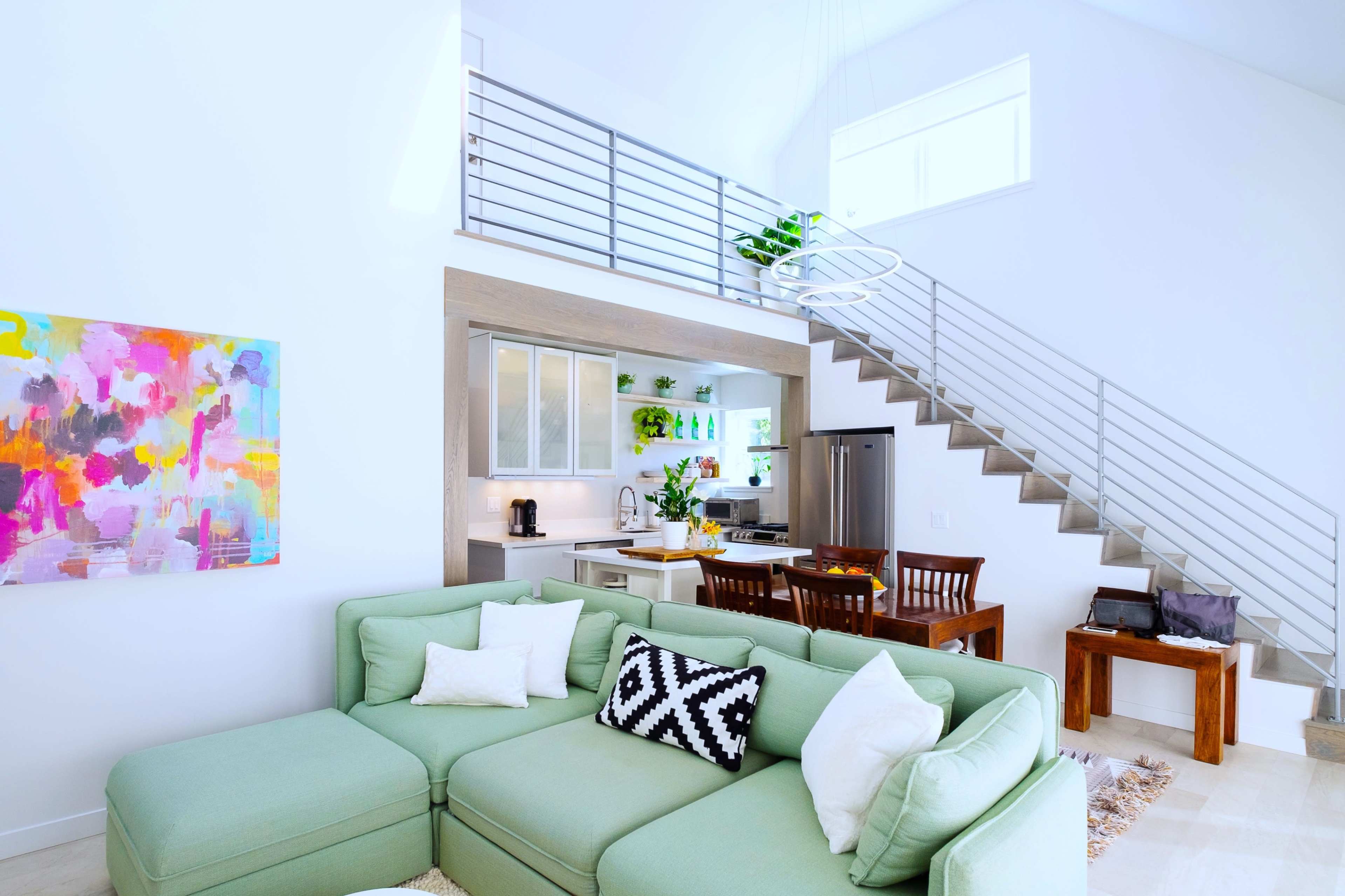 A bright living room features a large green sectional sofa, an abstract painting on the wall, and a dining area visible in the background.