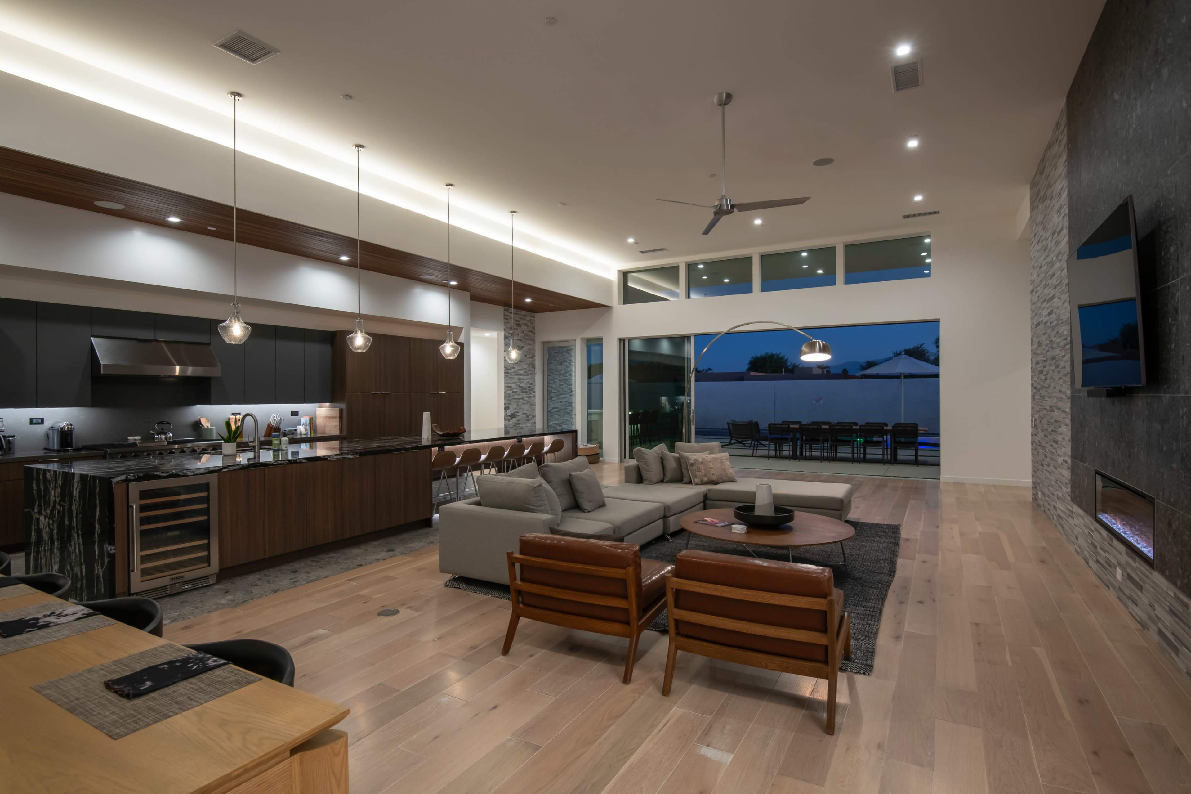 The image shows a modern open-concept living space featuring a kitchen, dining area, and lounge with large windows and contemporary furnishings.