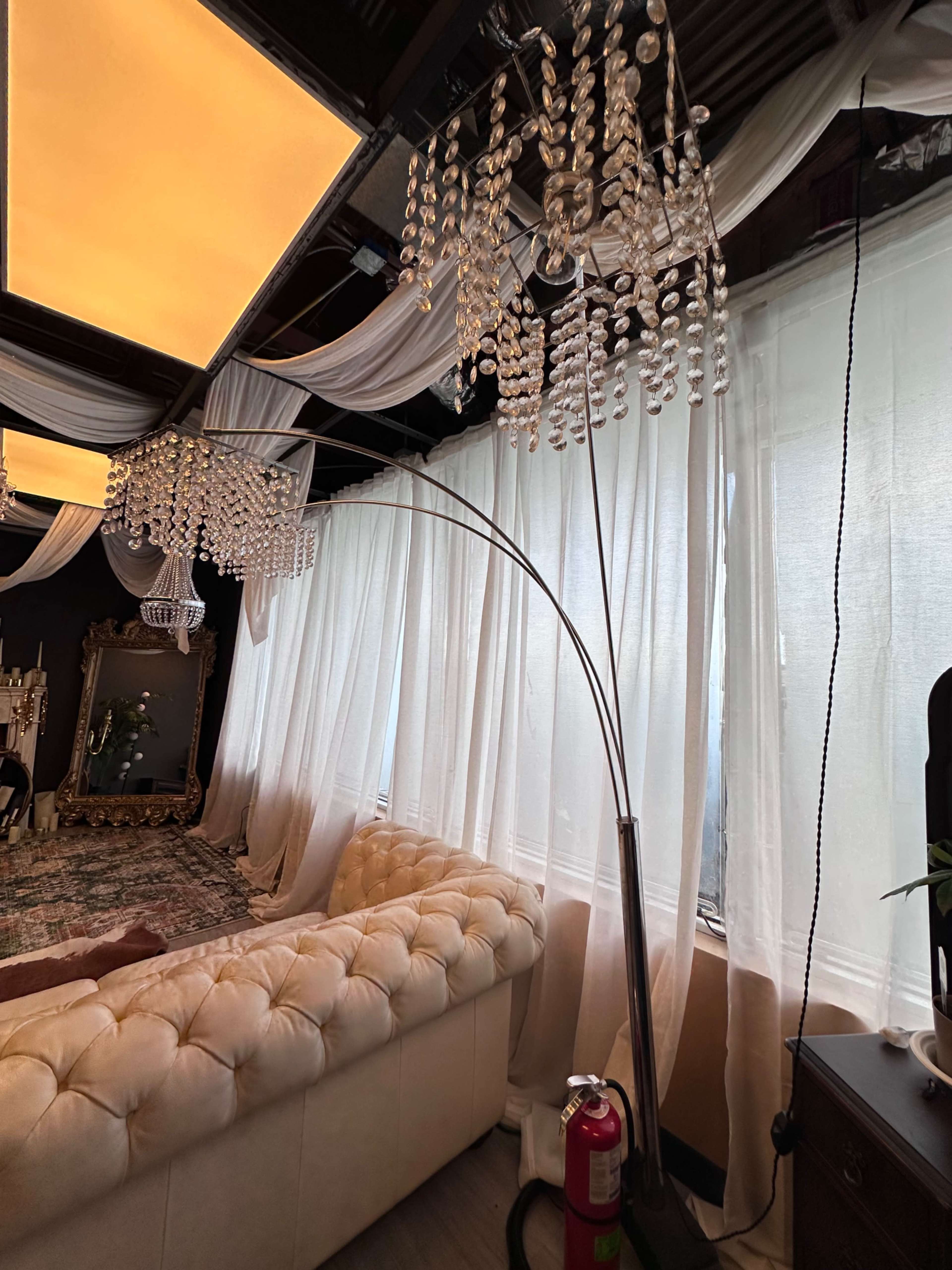 The image shows a stylish indoor space featuring a white tufted sofa, elegant chandeliers made of beads, sheer curtains, and a modern floor lamp.