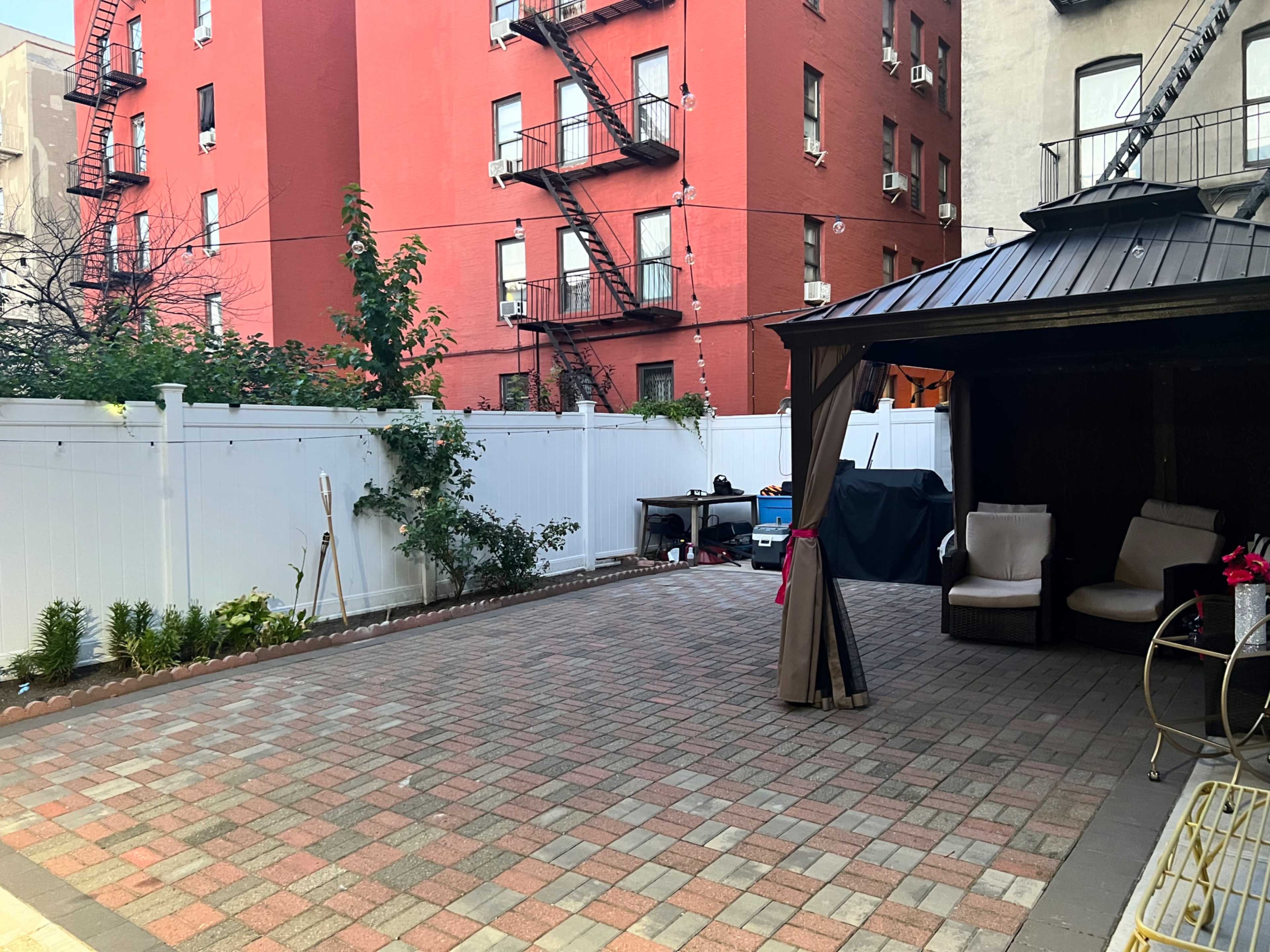 A paved backyard area with a gazebo and seating, surrounded by a white fence and red brick buildings in the background.