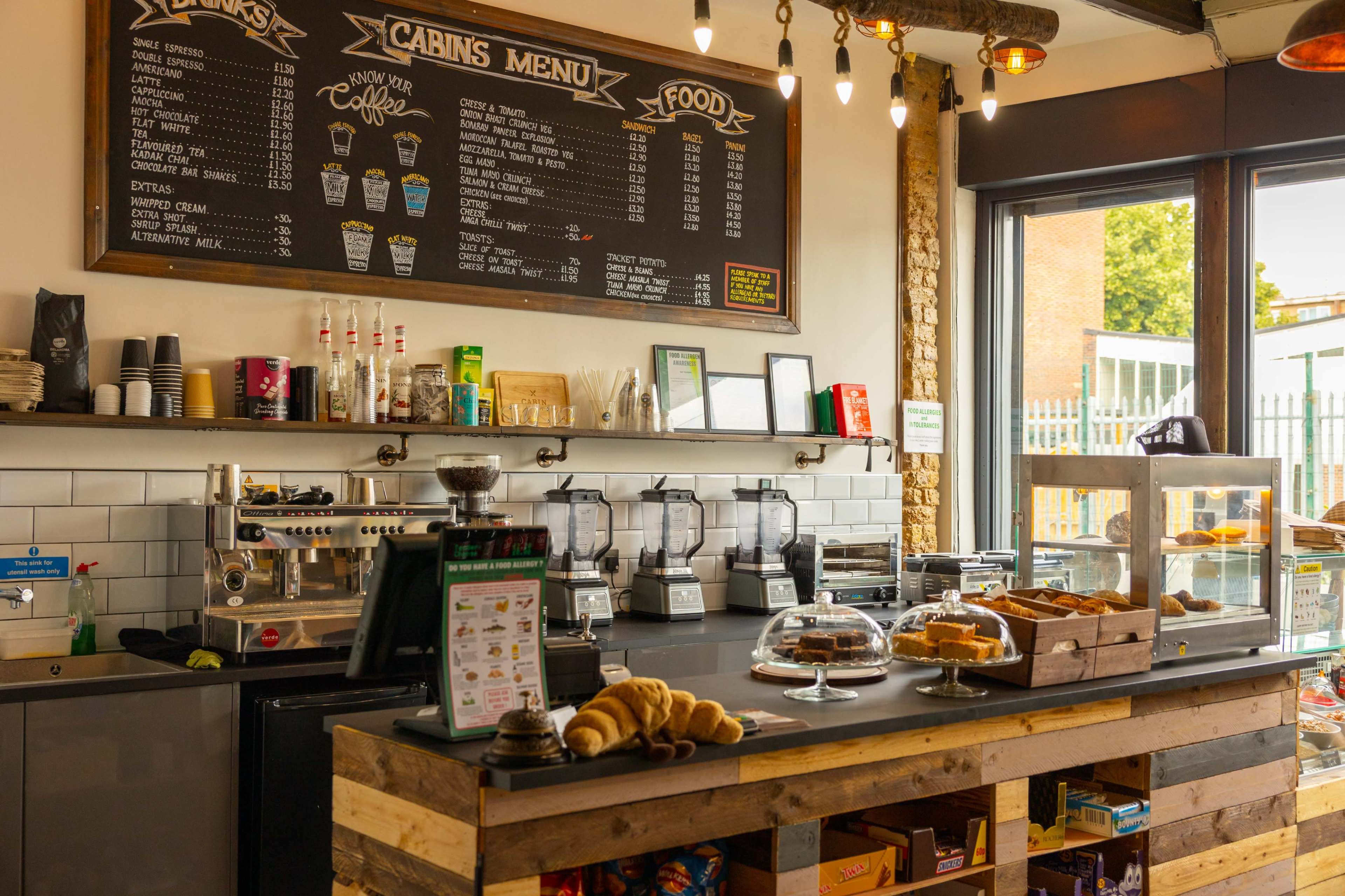 A cozy café interior featuring a menu board, a coffee machine, glass display cases with baked goods, and a rustic wooden counter.