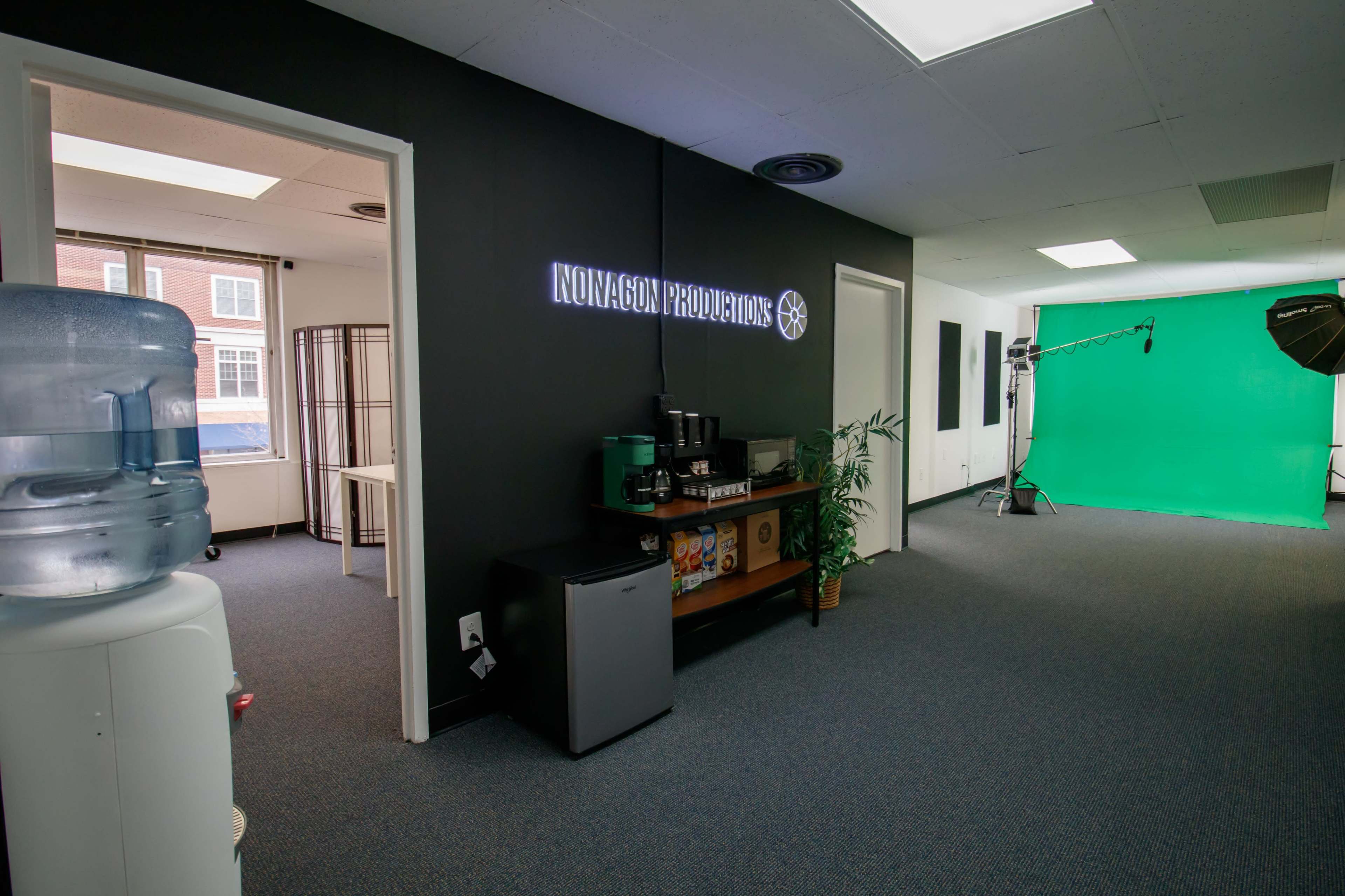 An office space with a black wall labeled "Innovagon Productions," a kitchenette area with a water cooler and coffee machine, and a green screen setup for video production.