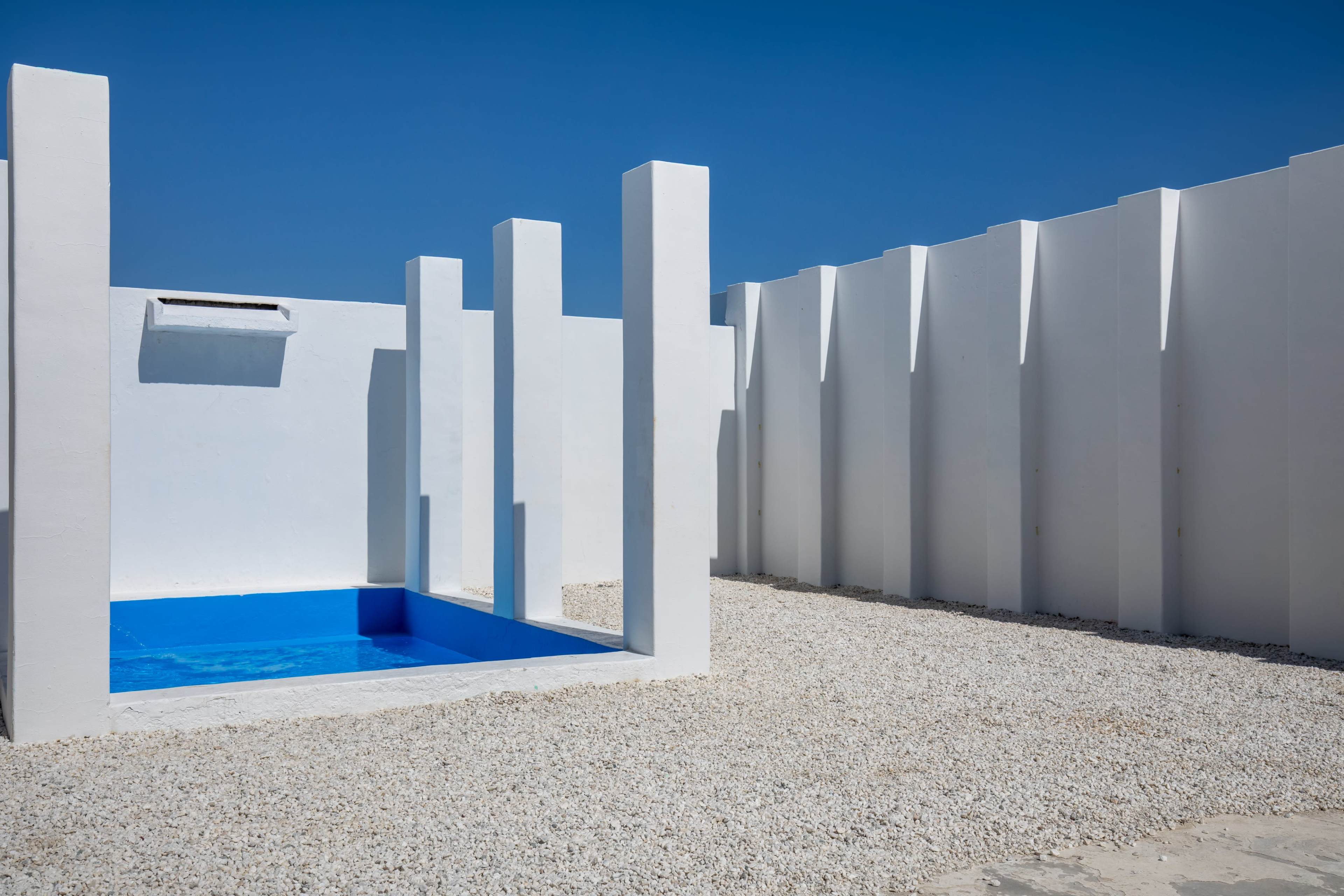 The image shows a modern, minimalistic outdoor space with a small blue pool surrounded by white walls and tall white columns.