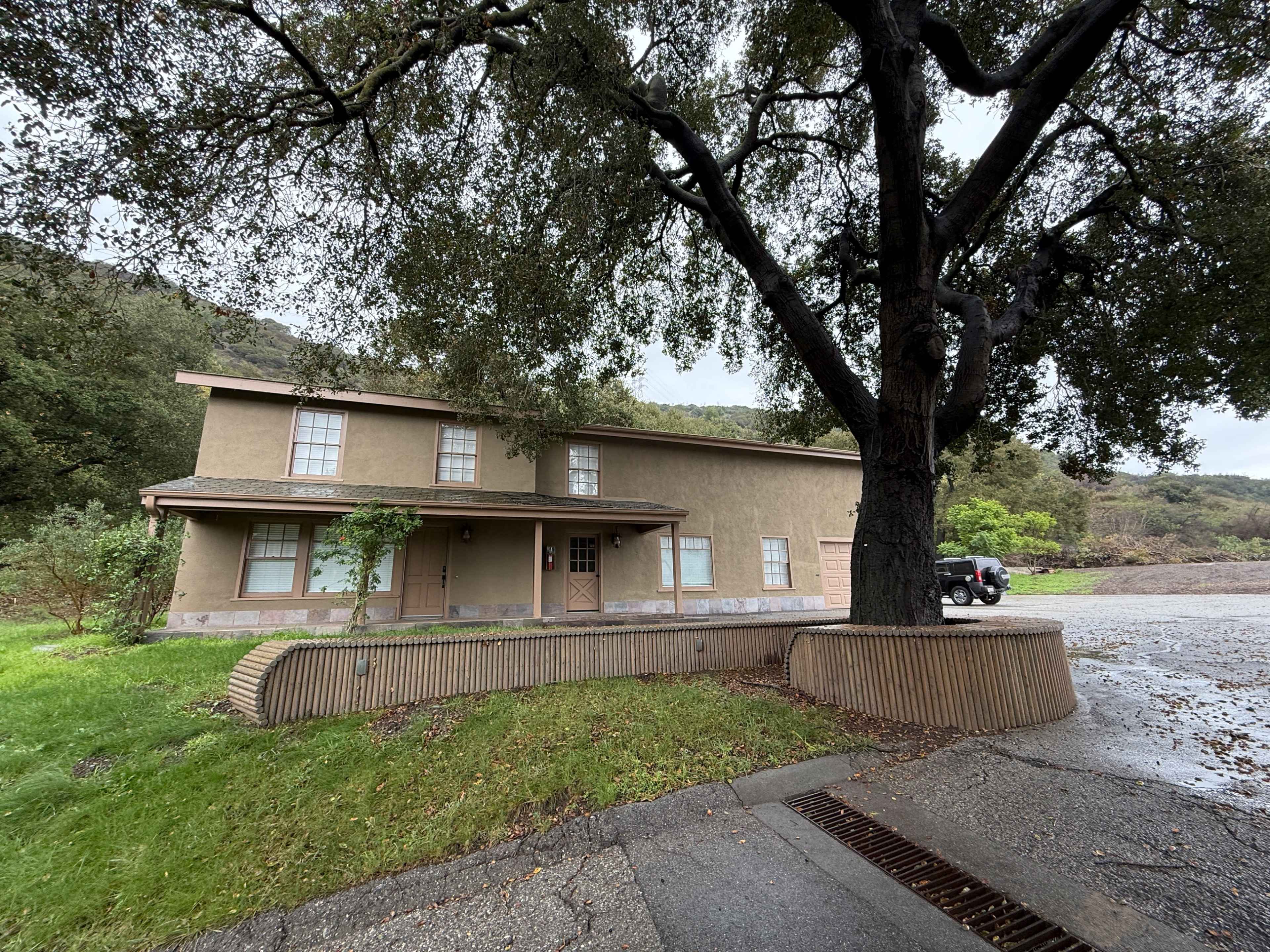 A two-story, beige building with a flat roof is set against a green landscape and a large tree on one side.