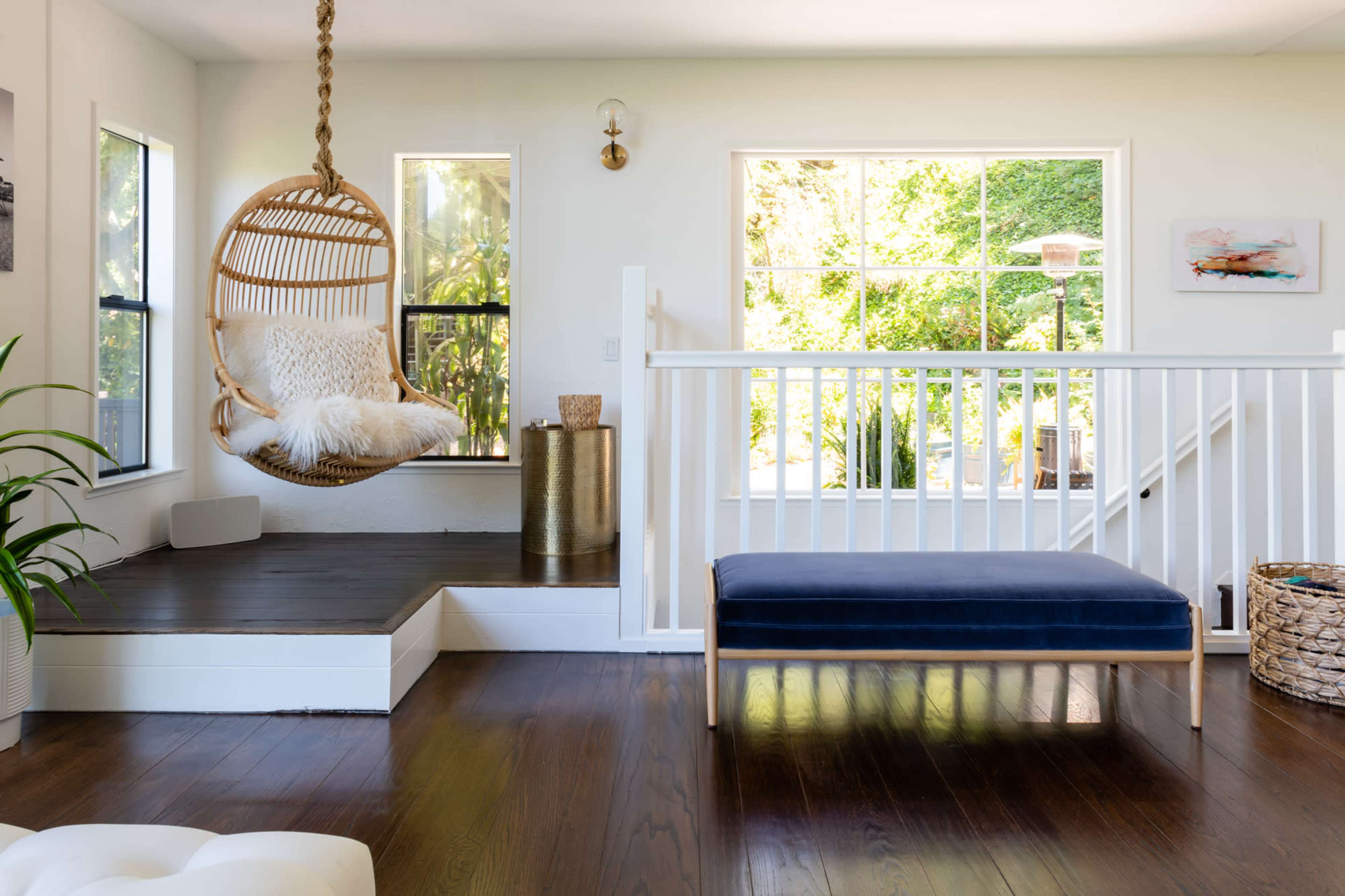The image shows a bright interior space featuring a hanging rattan chair beside a wooden platform, a navy bench, and large windows that offer a view of greenery outside.
