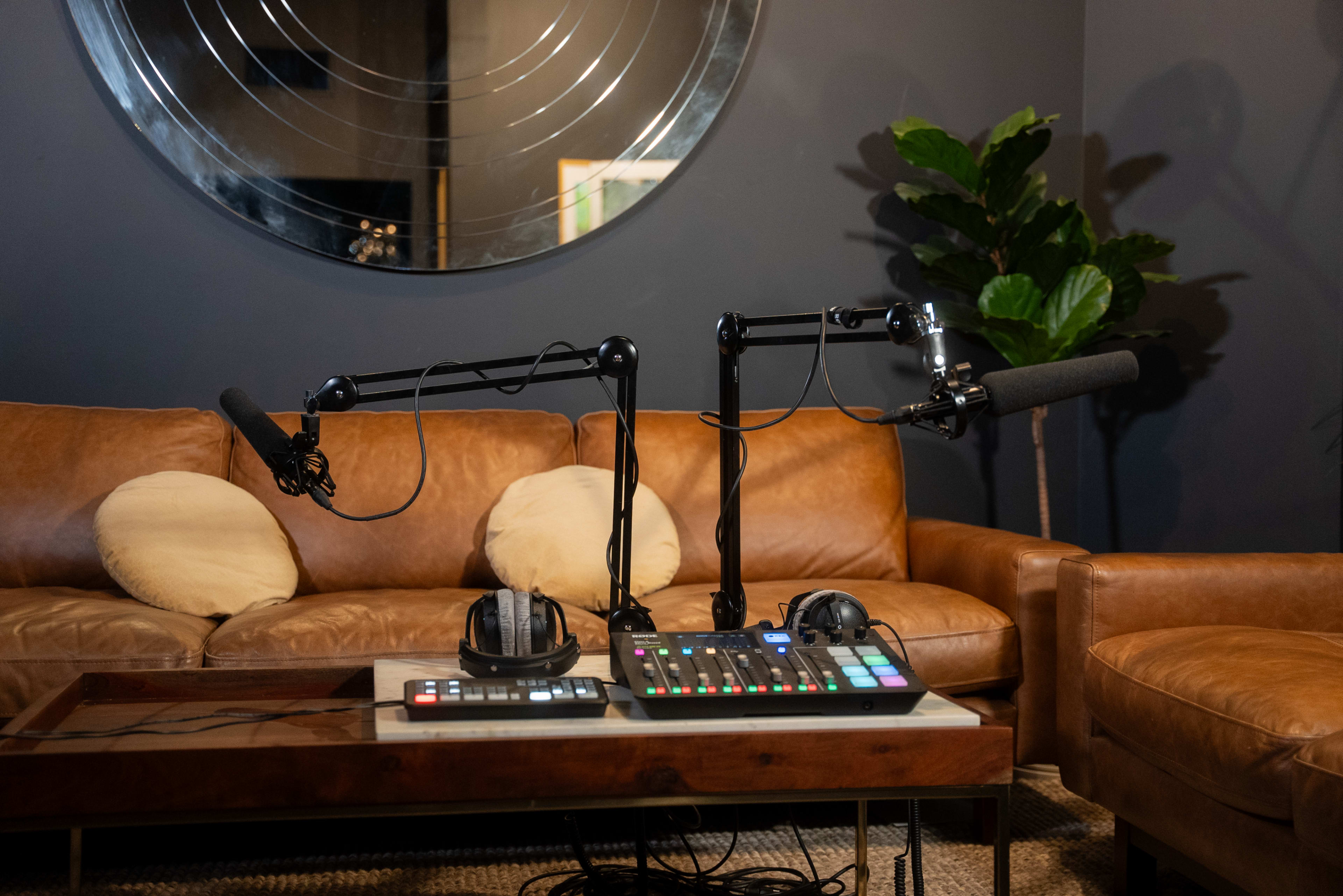 A recording setup featuring two microphone arms, a soundboard, and headphones is positioned on a coffee table in front of a brown leather sofa.