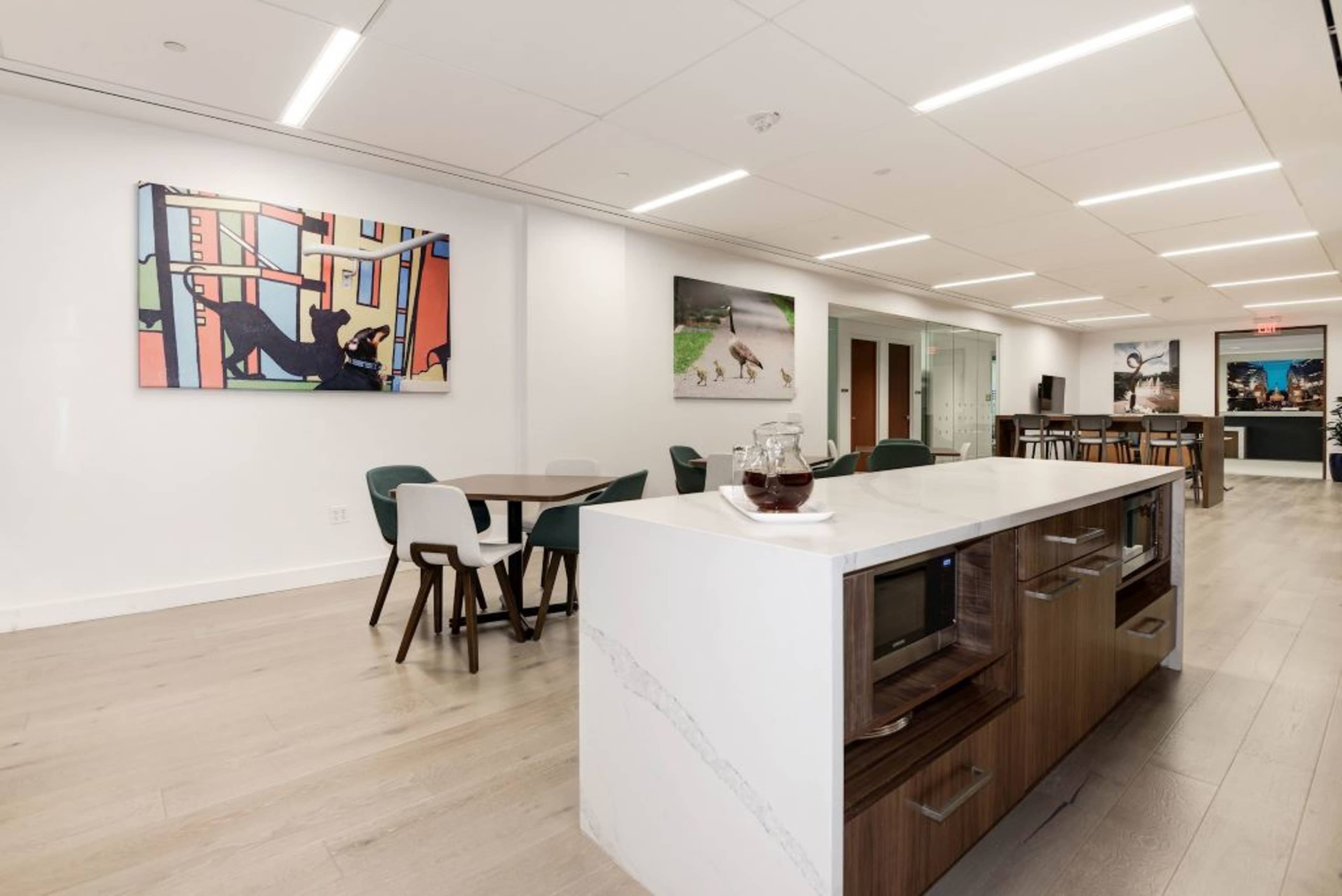 A modern office space with a kitchen area featuring a large countertop, microwaves, and dining tables arranged around the room, decorated with colorful artwork on the walls.