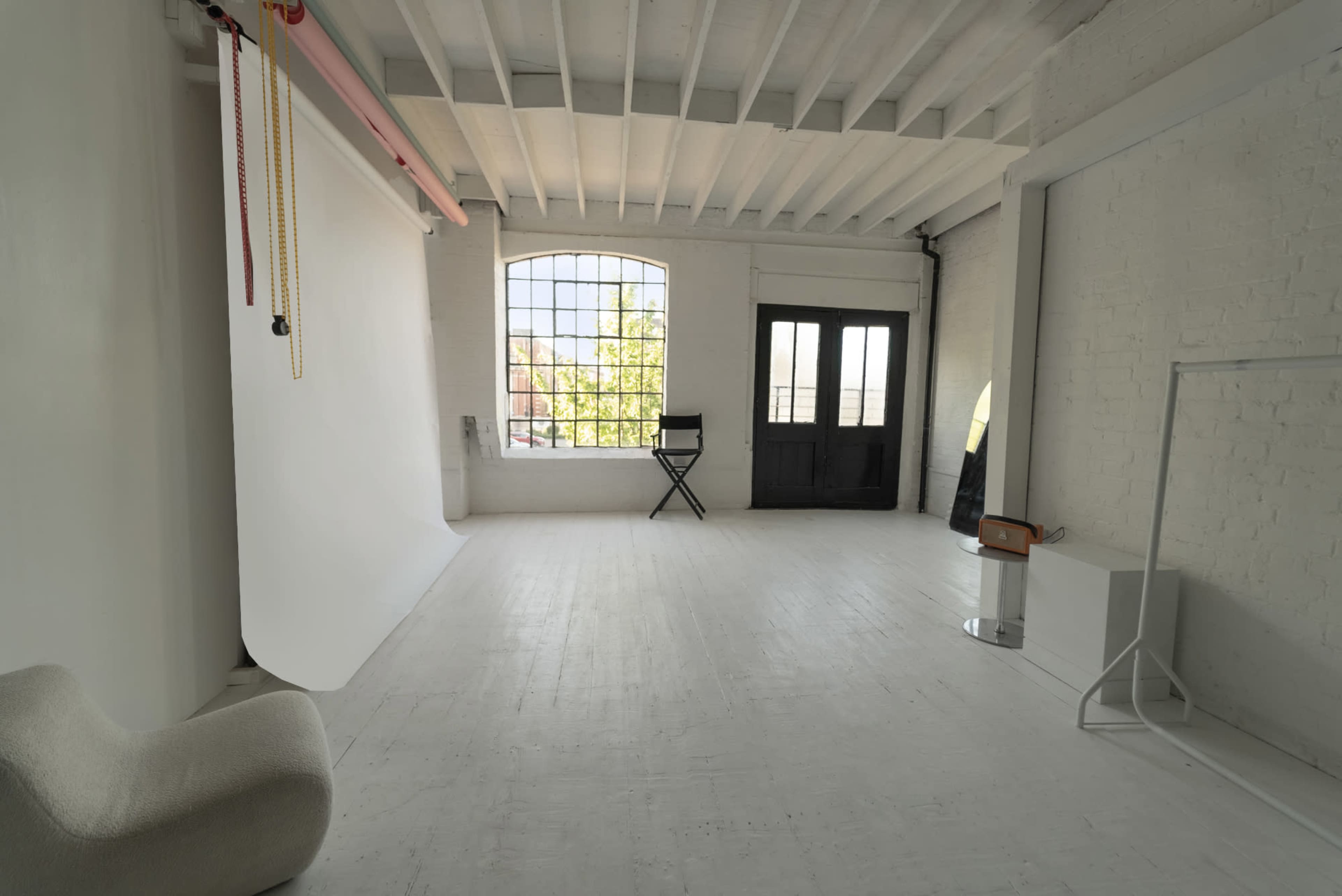 The image shows a spacious, well-lit interior with white walls, wooden beams, a black chair, and large windows.