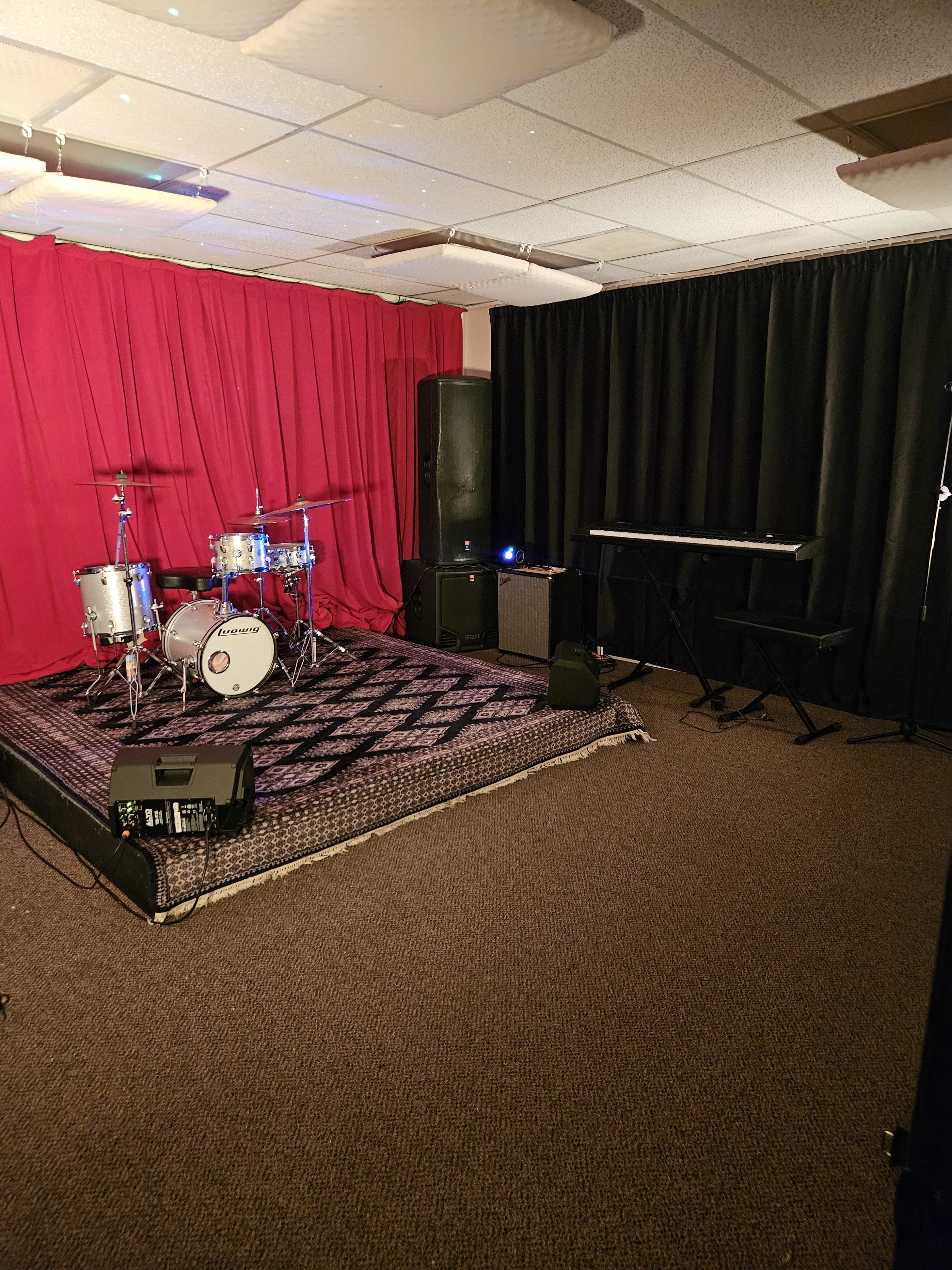 The image shows a music rehearsal space featuring a drum set, a keyboard, and a red curtain on one wall, with black curtains and carpet flooring.