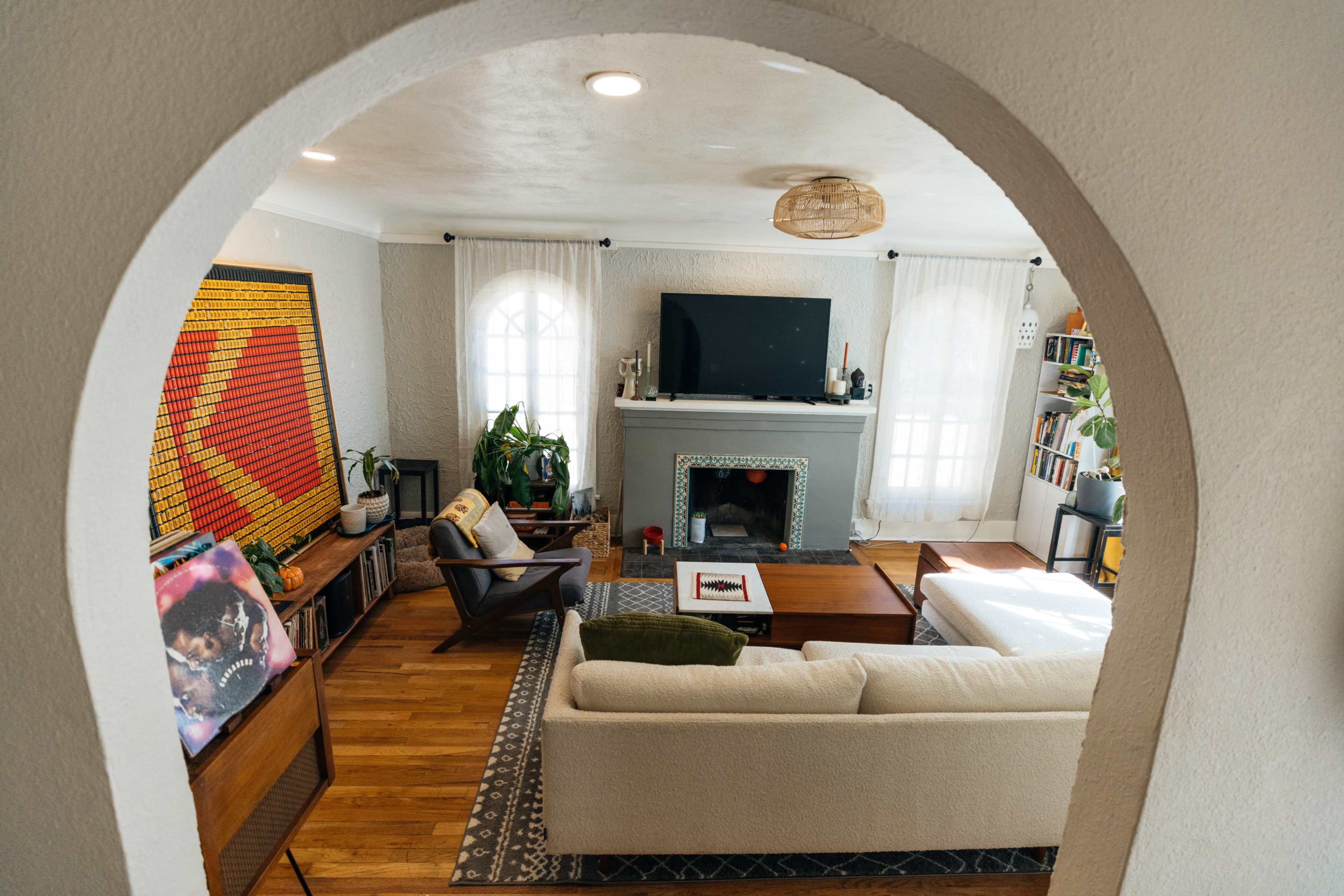 The image shows a cozy living room featuring a television above a fireplace, wooden furniture, plants, and artwork on the walls.