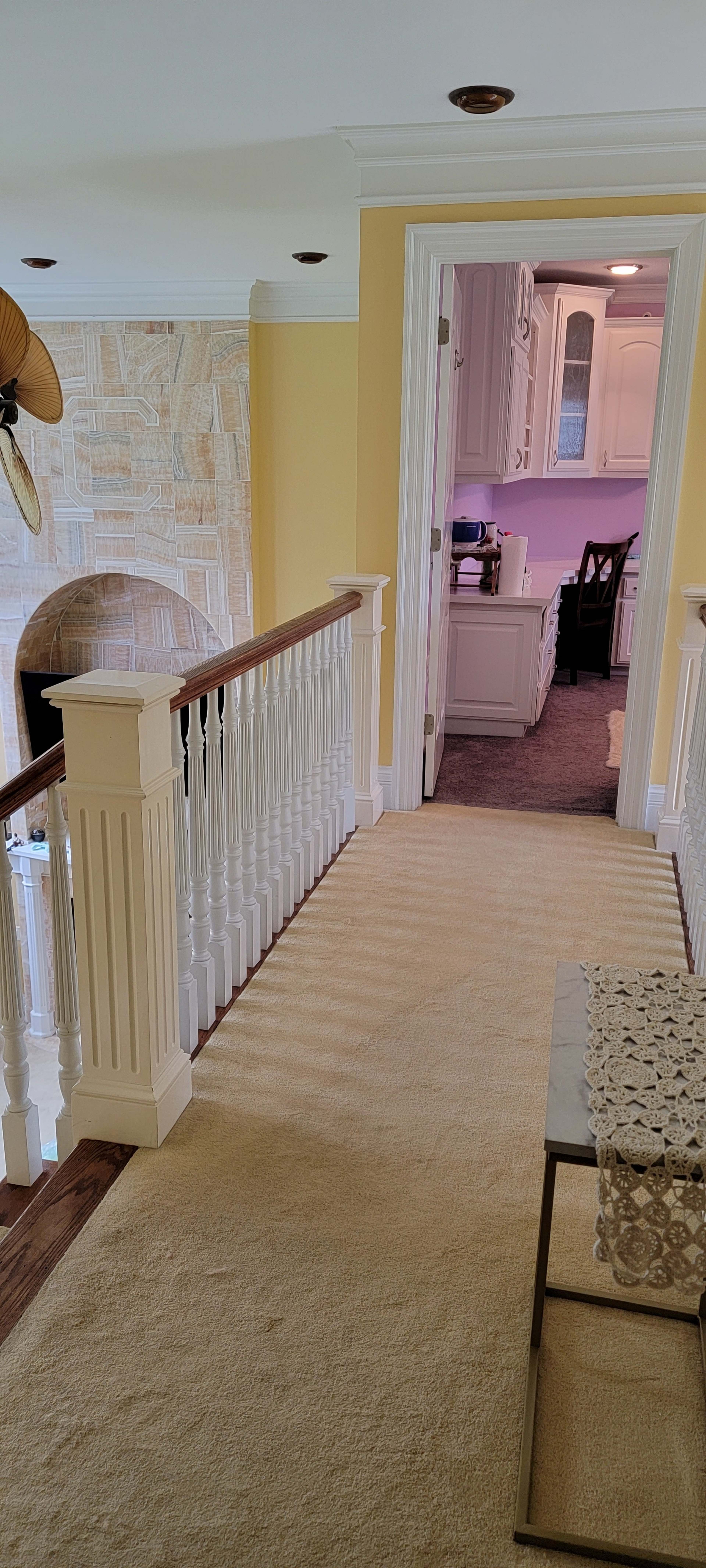 A carpeted hallway with a white railing leading to a brightly lit kitchen in the background.