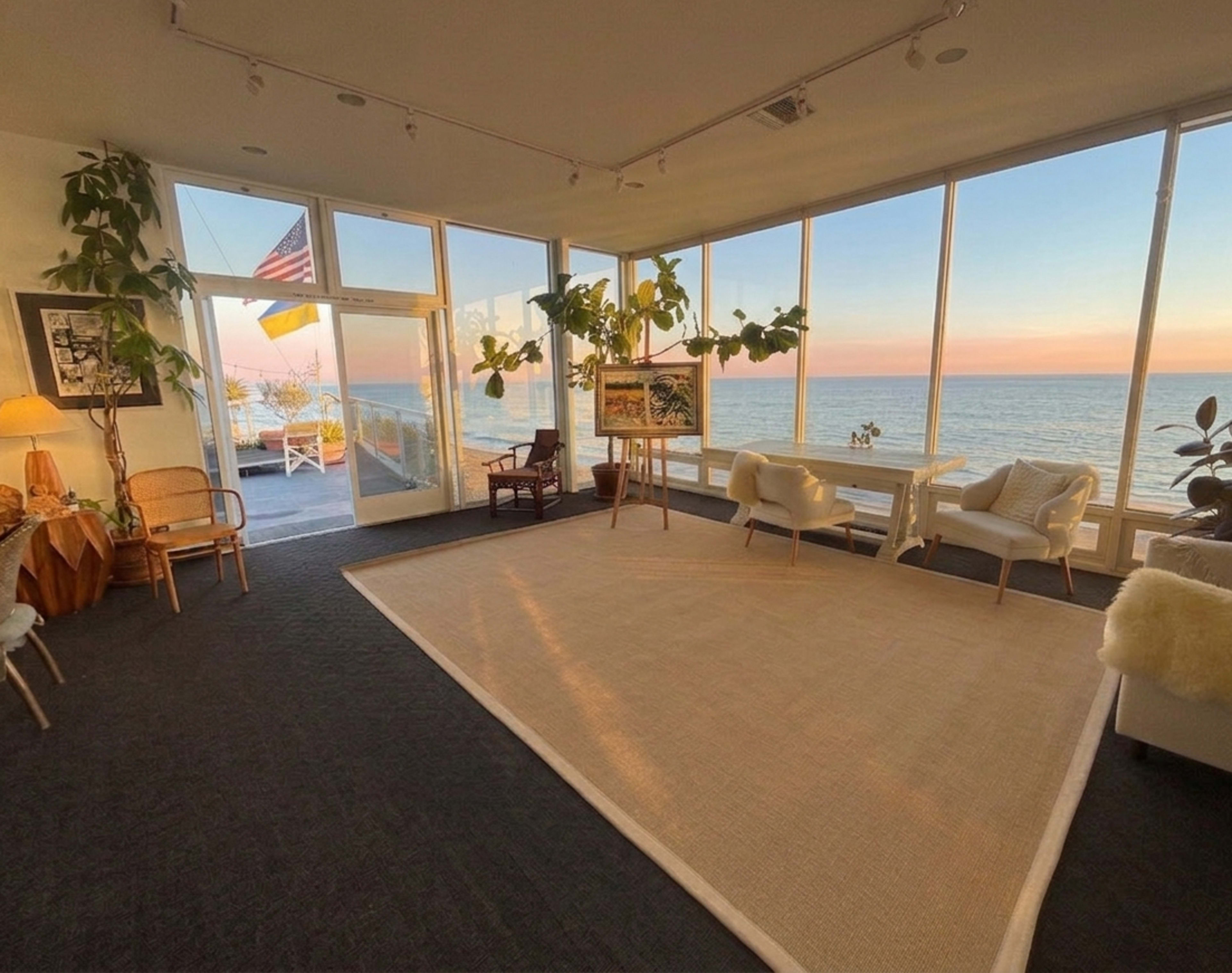 The image shows a spacious, modern living area with large windows overlooking the ocean, featuring several plants, furniture, and a light-colored rug.