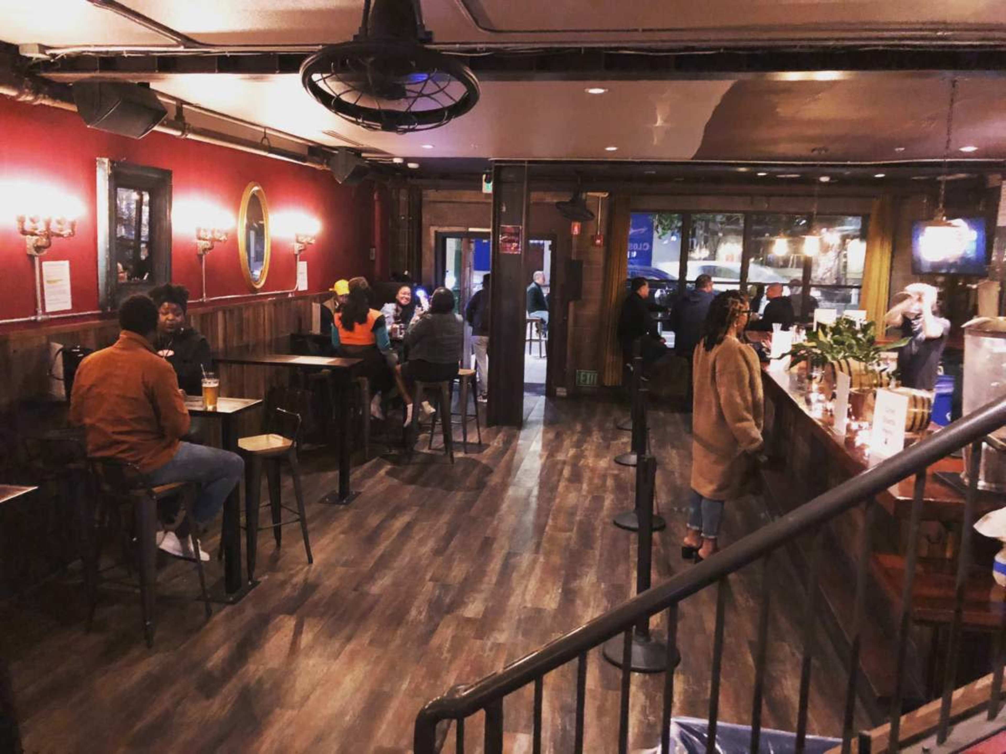 The image shows a dimly lit bar with patrons seated at high tables and the bar counter, surrounded by red walls and warm lighting.