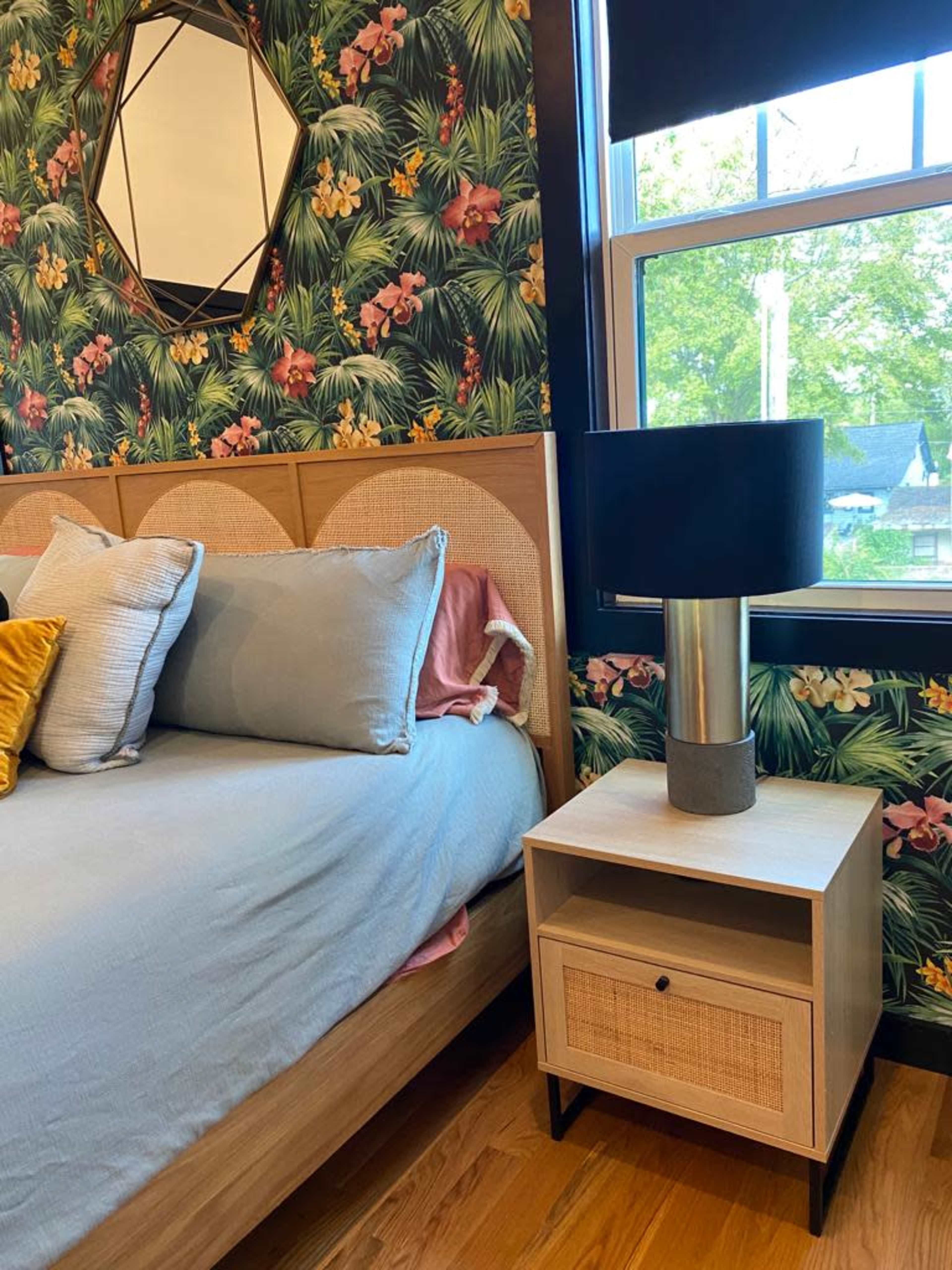A bedroom featuring a bed with light blue bedding, a bedside table with a black lamp, and a wall adorned with floral wallpaper.