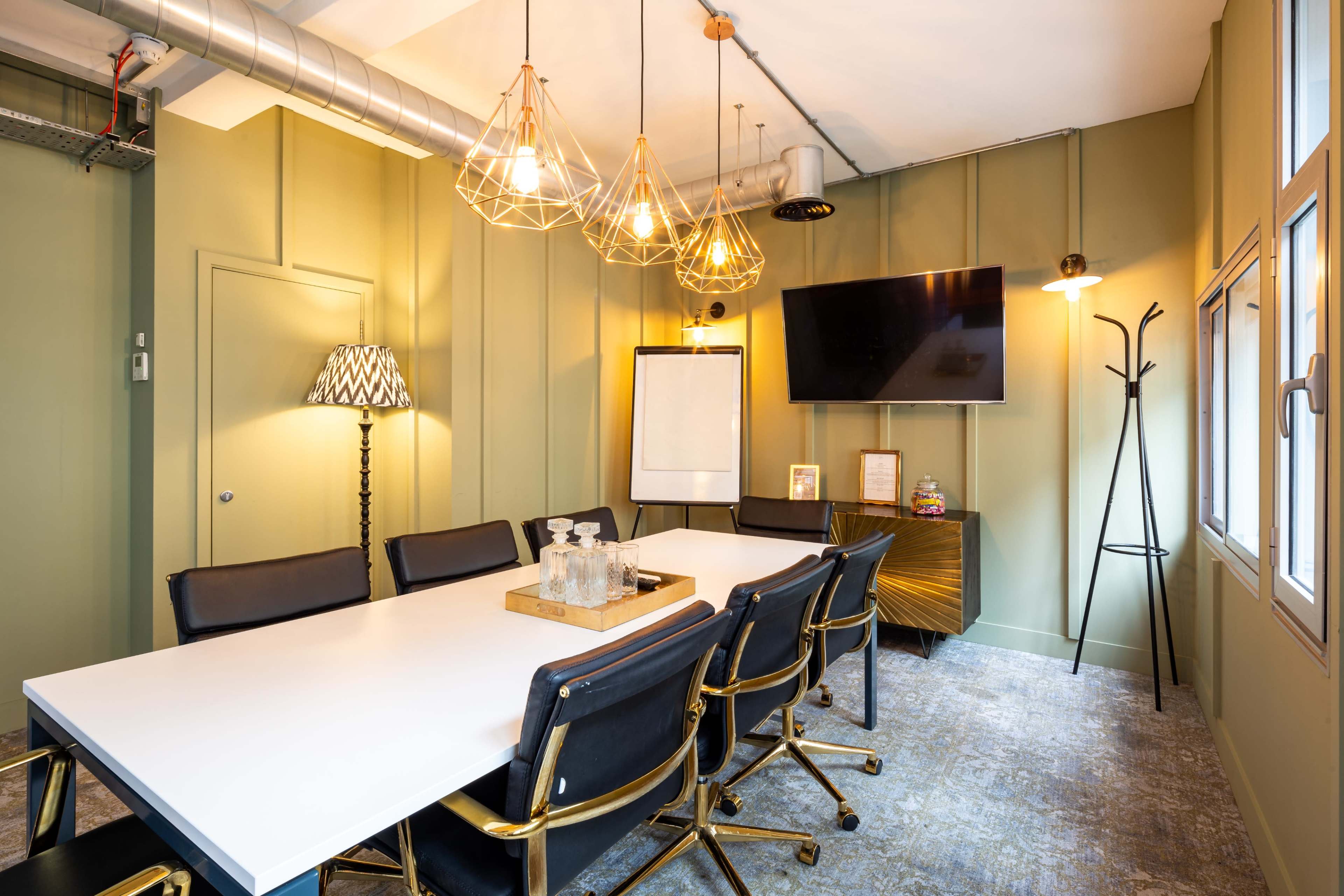 A conference room features a rectangular white table surrounded by black chairs, with pendant lights overhead and a large wall-mounted screen.