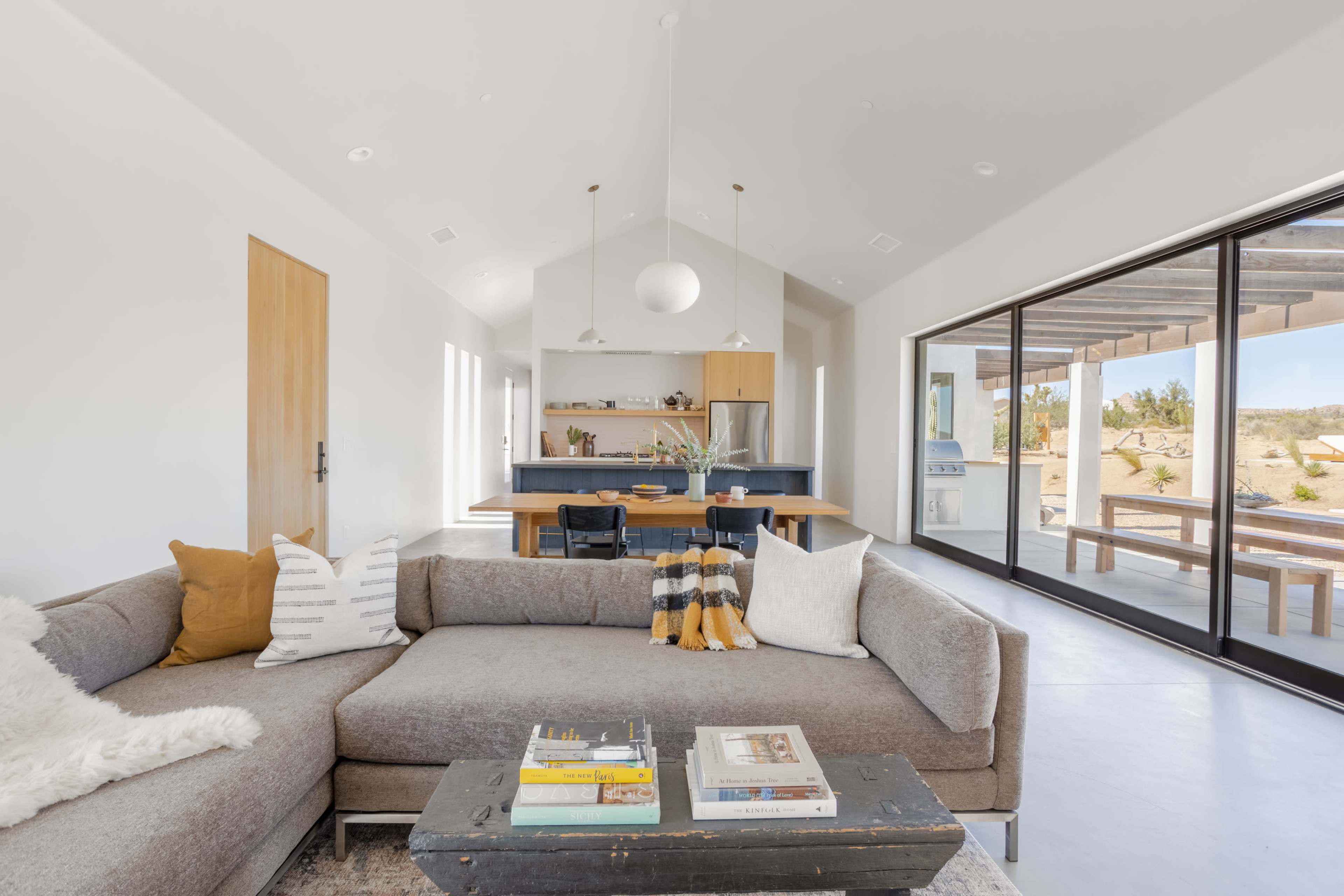 A spacious living area features a gray sectional sofa, a wooden dining table, and large sliding glass doors that open to an outdoor space.