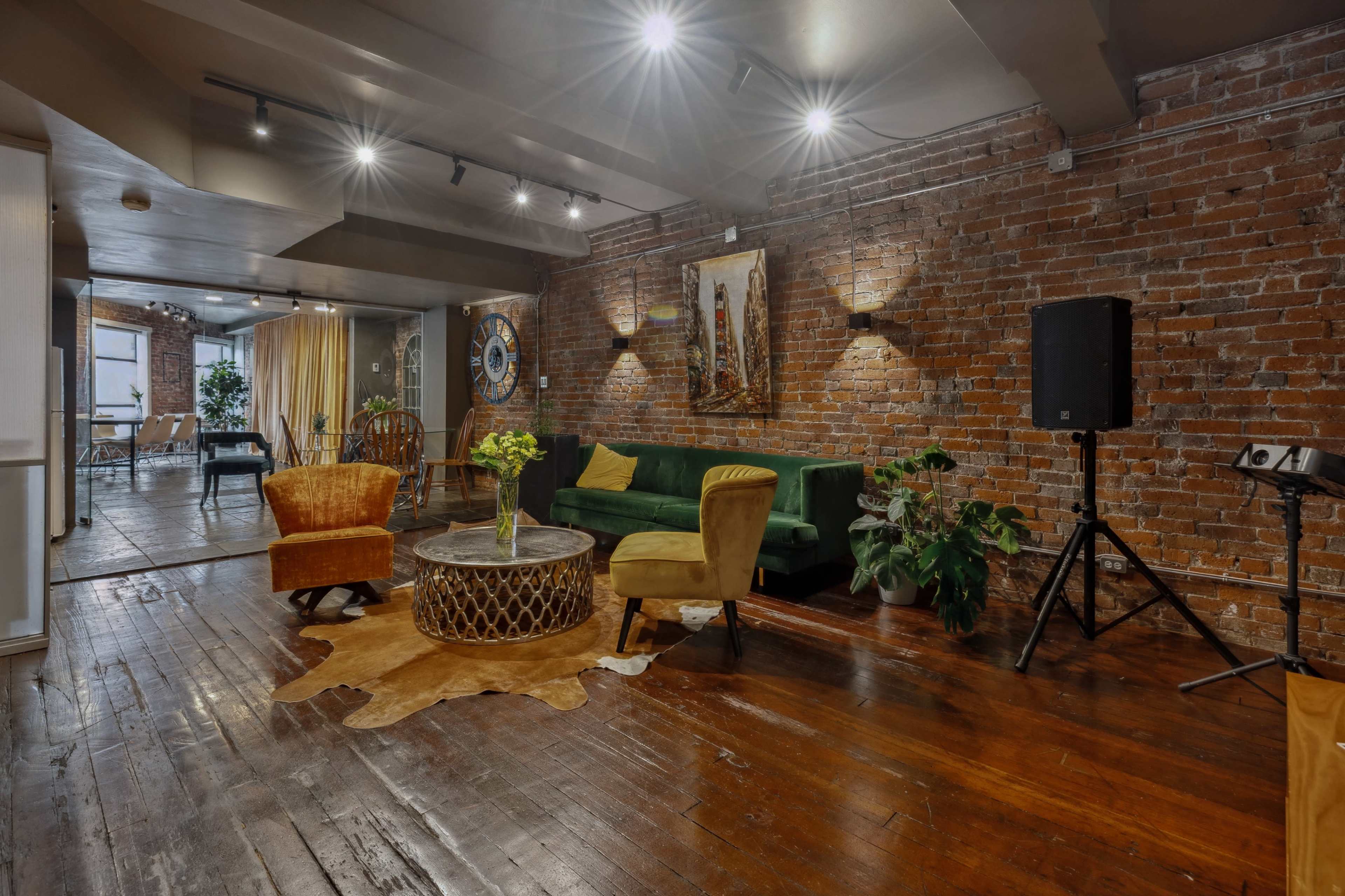 The image shows a spacious room with exposed brick walls, hardwood flooring, and a combination of seating areas, including chairs and a green couch.