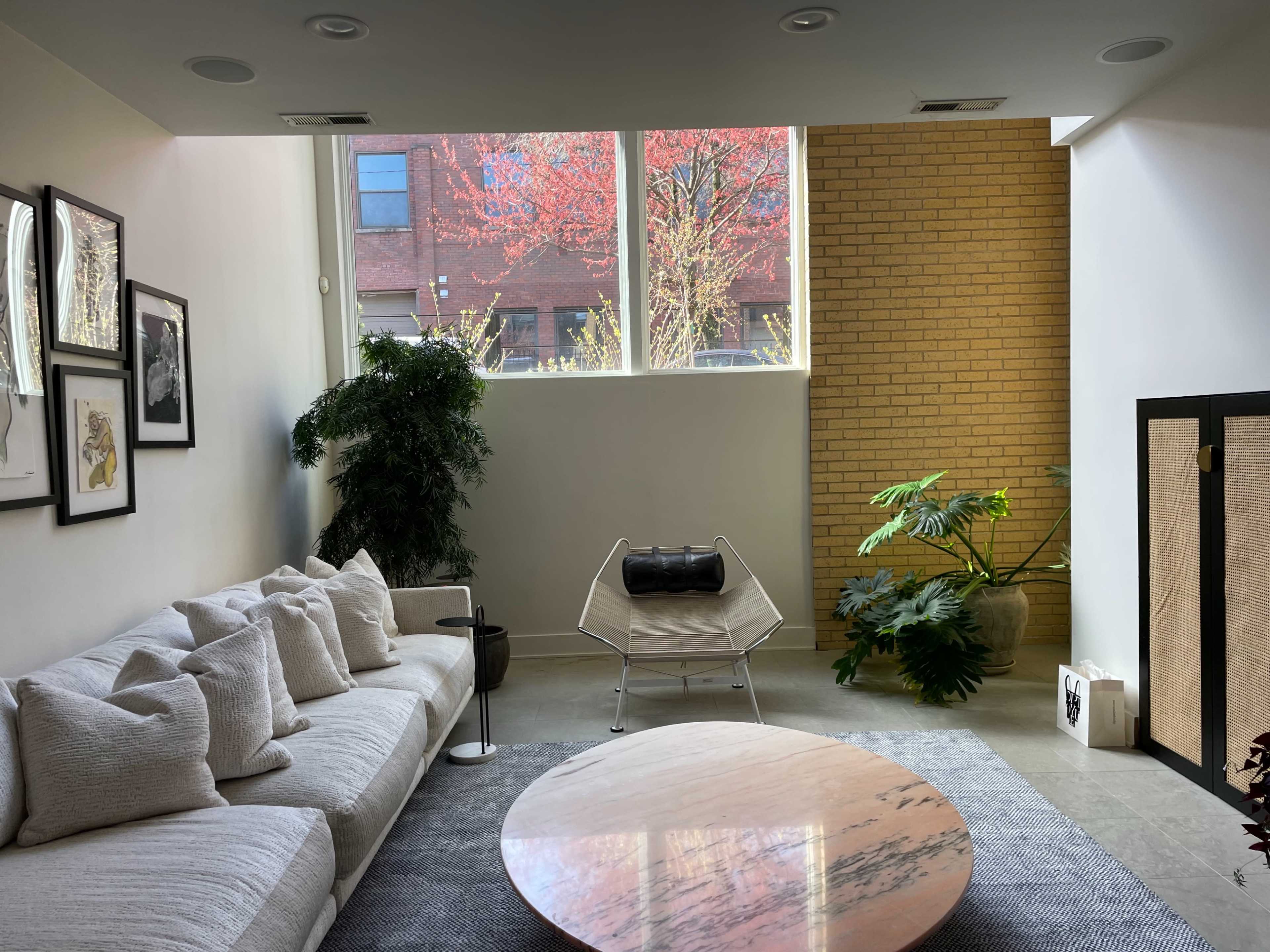The image depicts a modern living room featuring a white couch, a round marble coffee table, a potted plant, and large windows overlooking a brick wall with a view of trees outside.