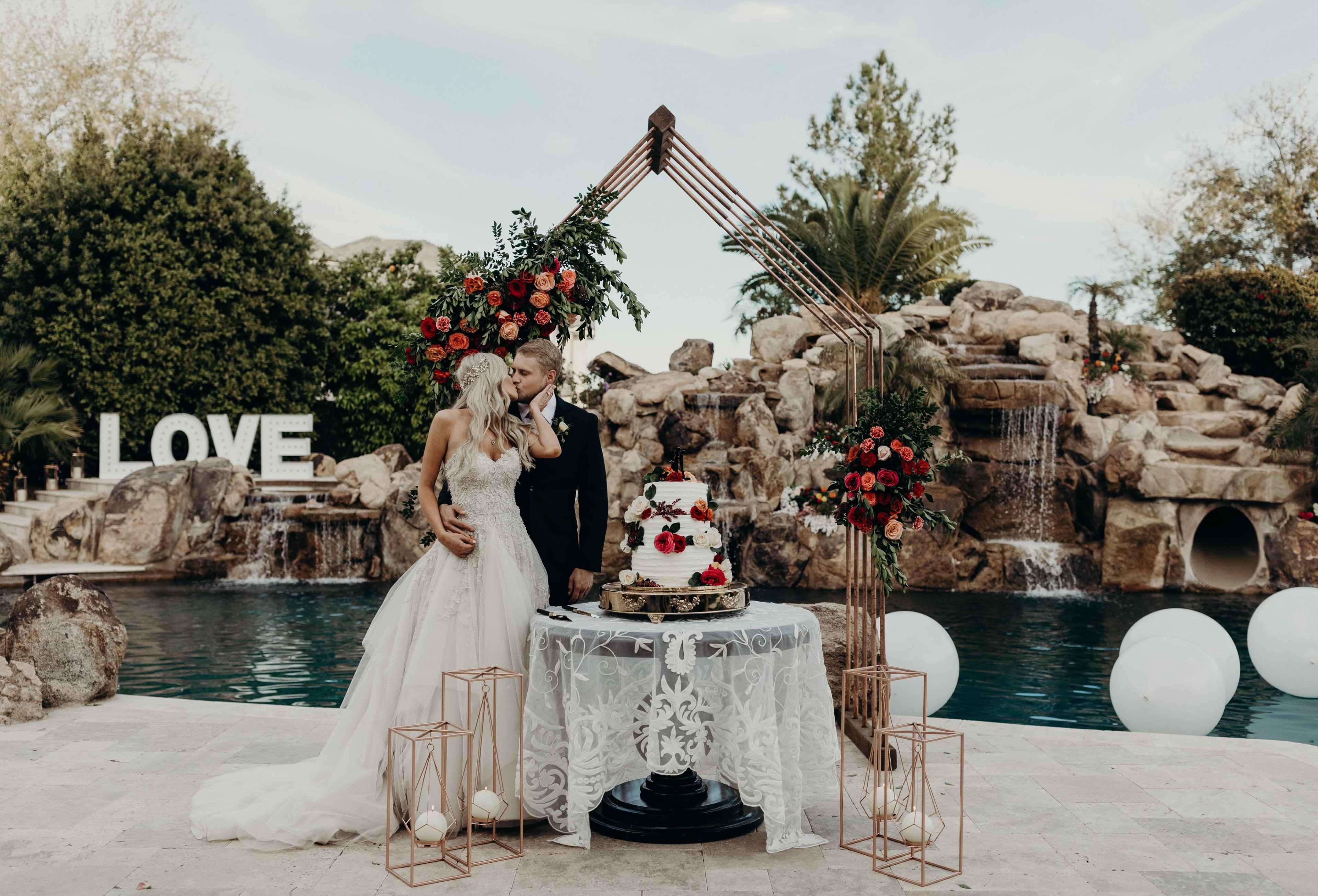 A couple stands together near a beautifully decorated wedding cake under an arch adorned with flowers, with a pool and stone waterfall in the background.