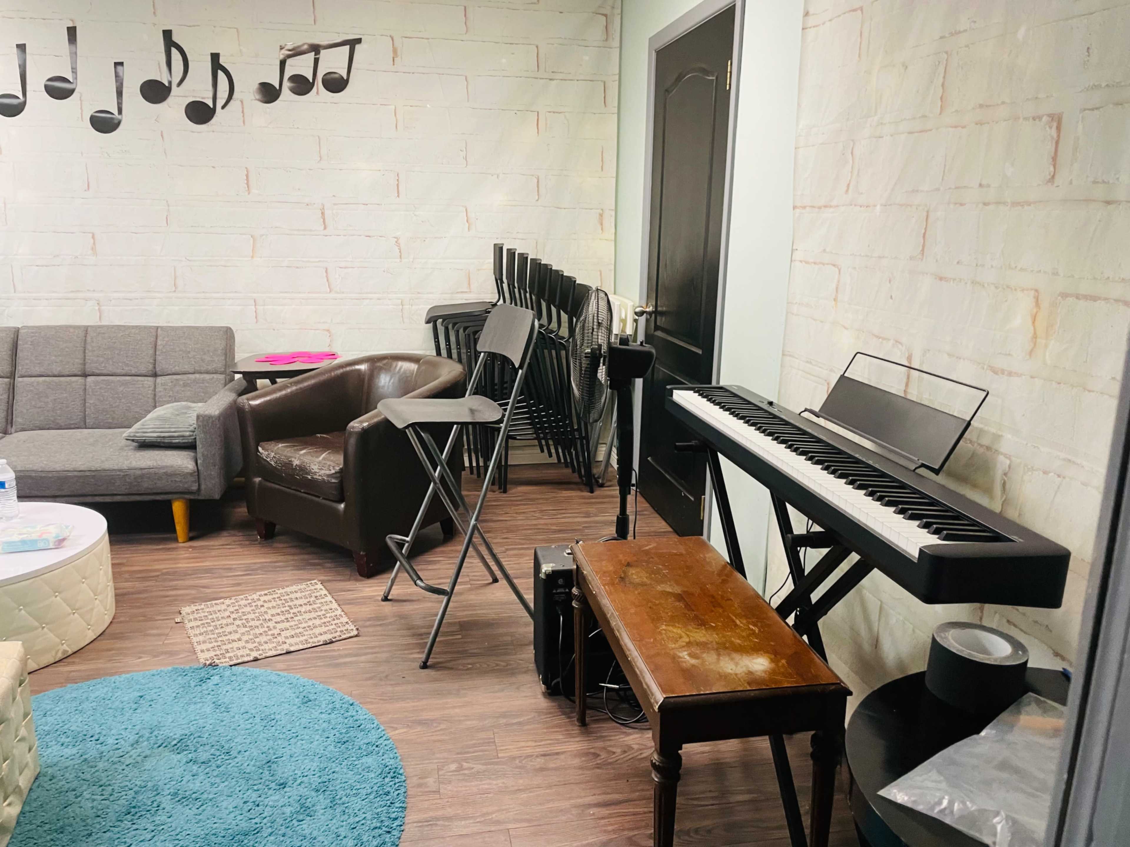 A room with a gray couch, a brown chair, a keyboard setup, and several stacked chairs against the wall.