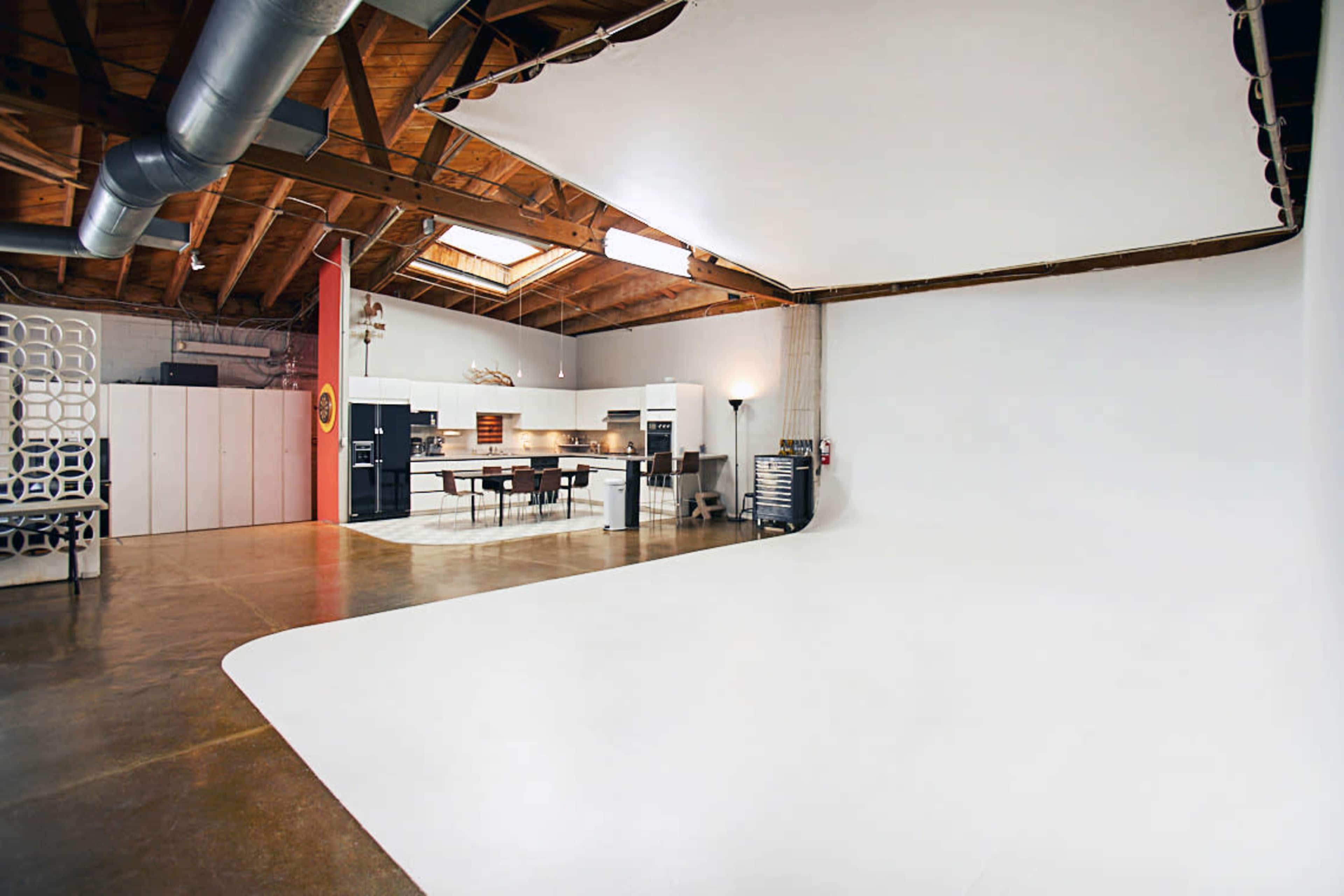 An industrial-style studio space with a large white backdrop, wooden beams, and a kitchen area in the background.