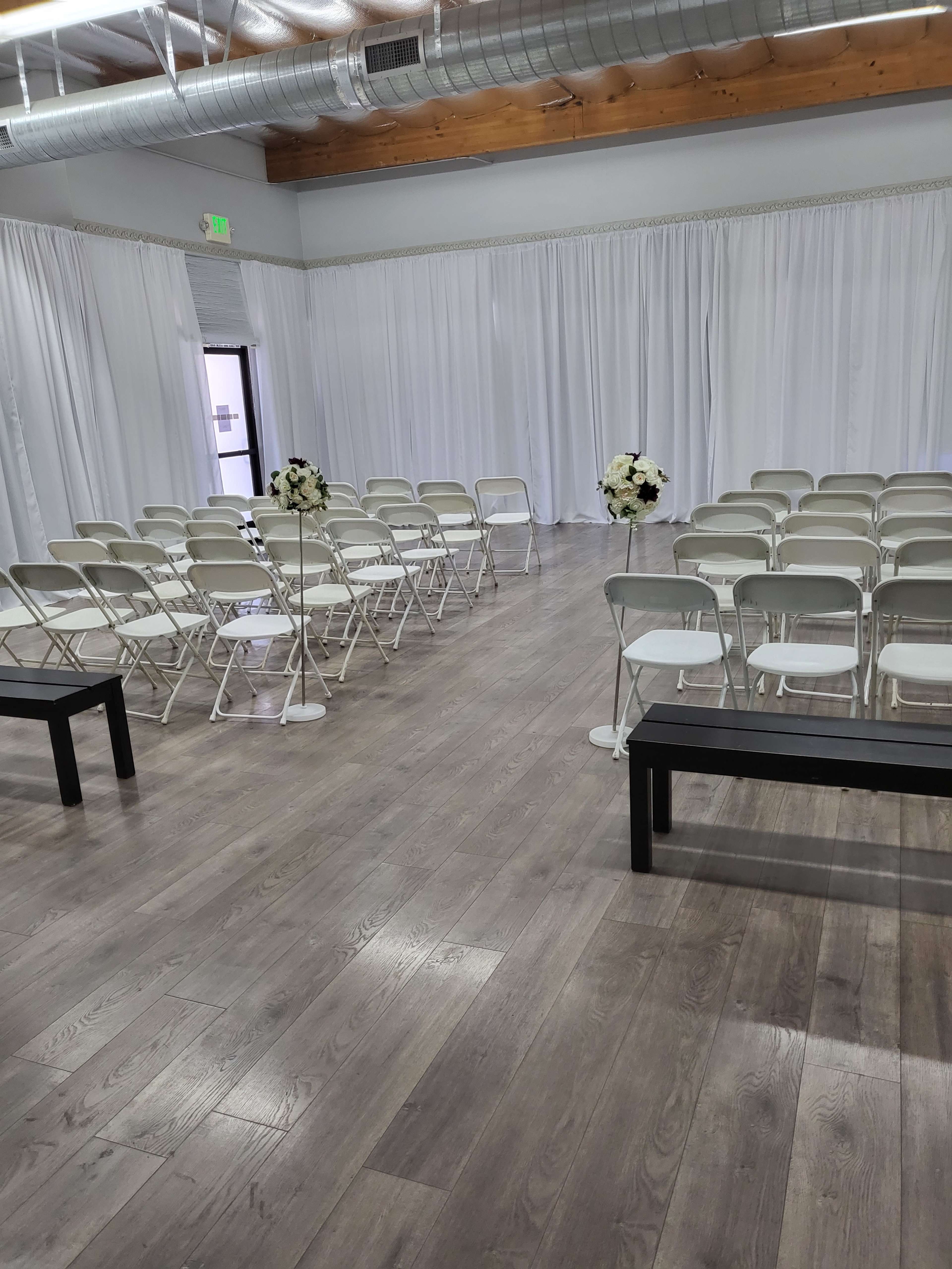 A spacious indoor venue set up for a ceremony, featuring rows of white folding chairs and flower arrangements on pedestals.