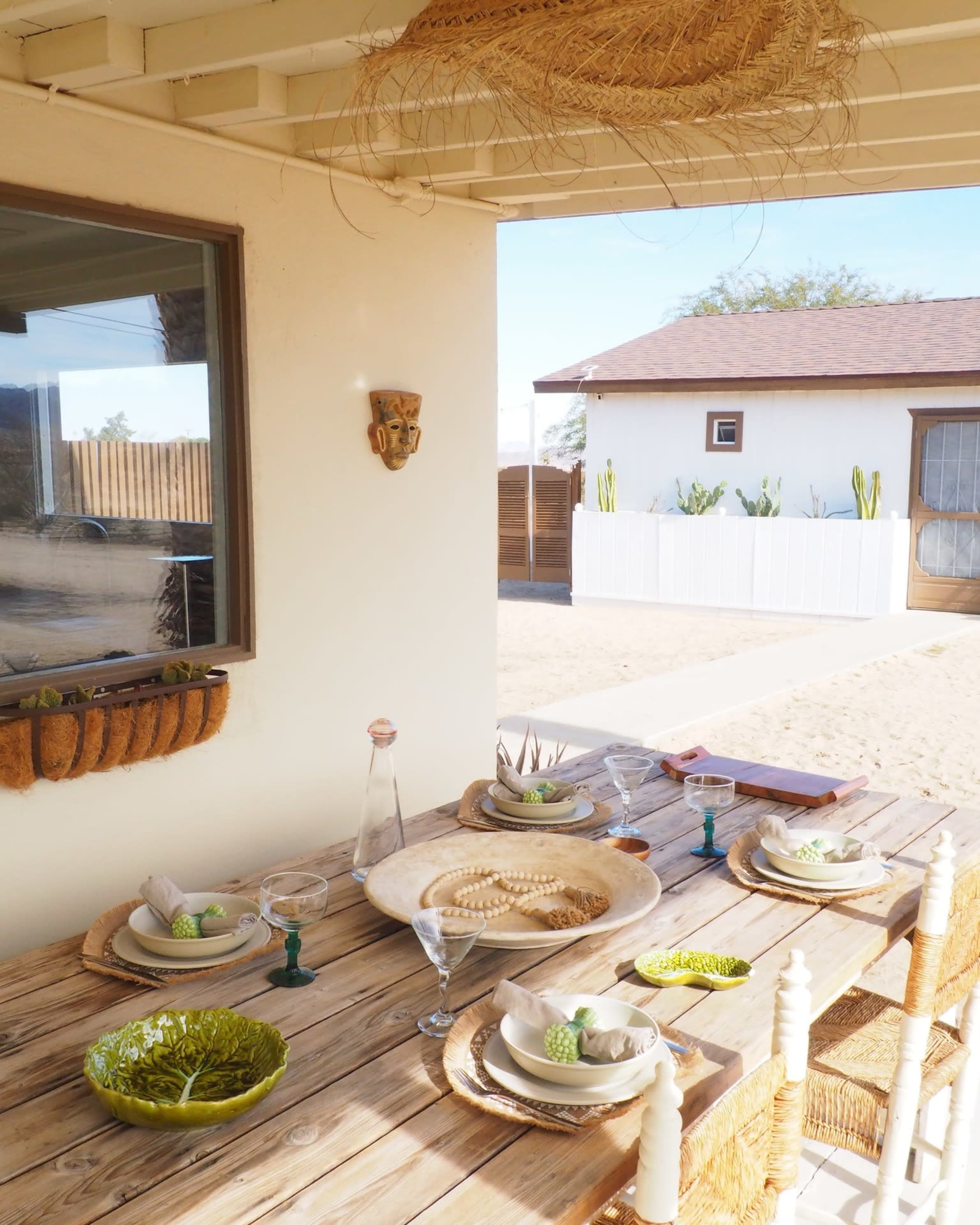 A wooden table is set with plates, glasses, and decorative items under a covered outdoor space, overlooking a sandy area and a white fence.