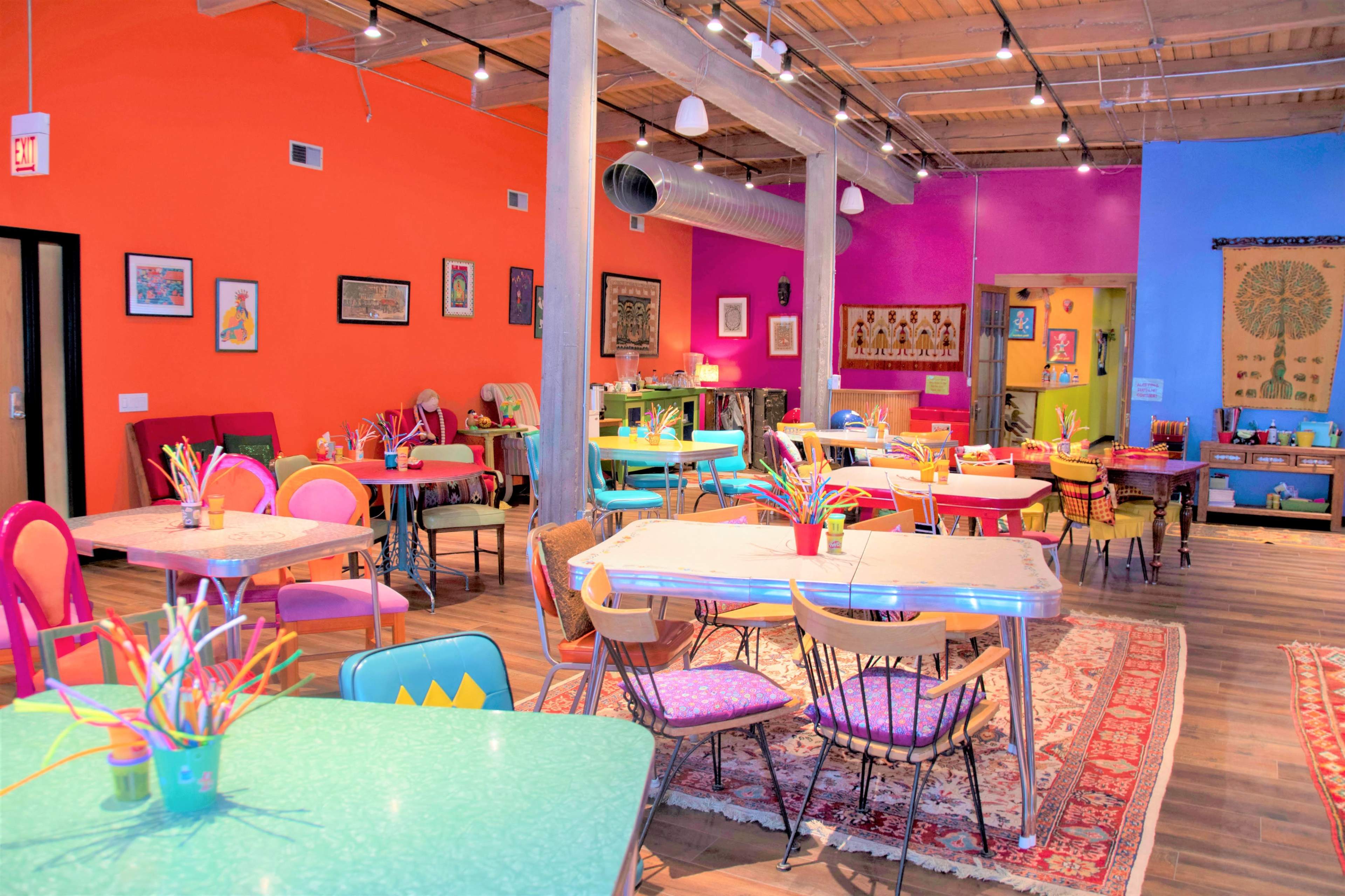 A vibrant cafe features colorful tables and chairs, craft supplies, and eclectic artwork on the walls.