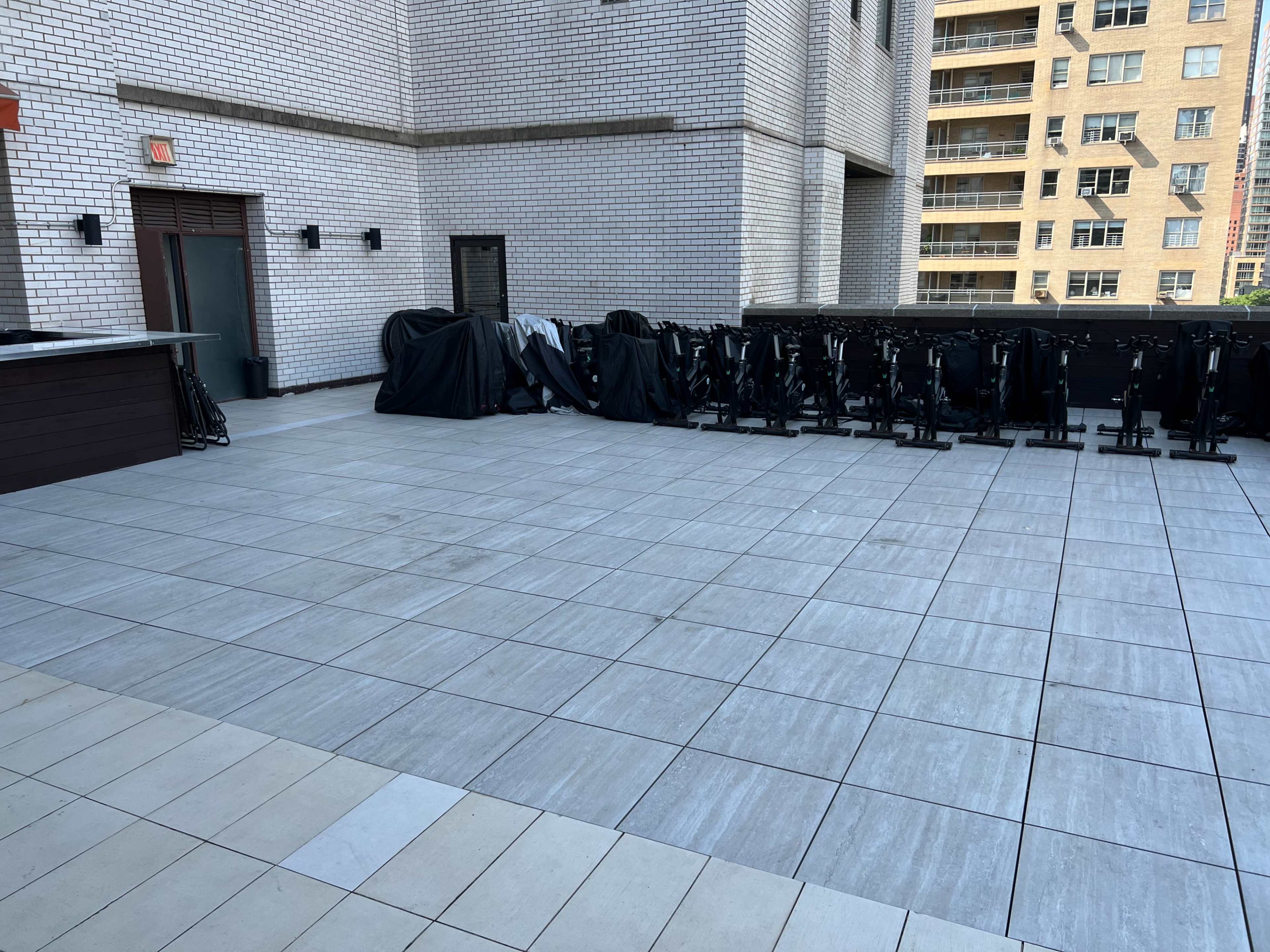 The image shows a rooftop terrace with gray tiled flooring, featuring a bar area and several stationary bicycles covered with black tarps against a wall.
