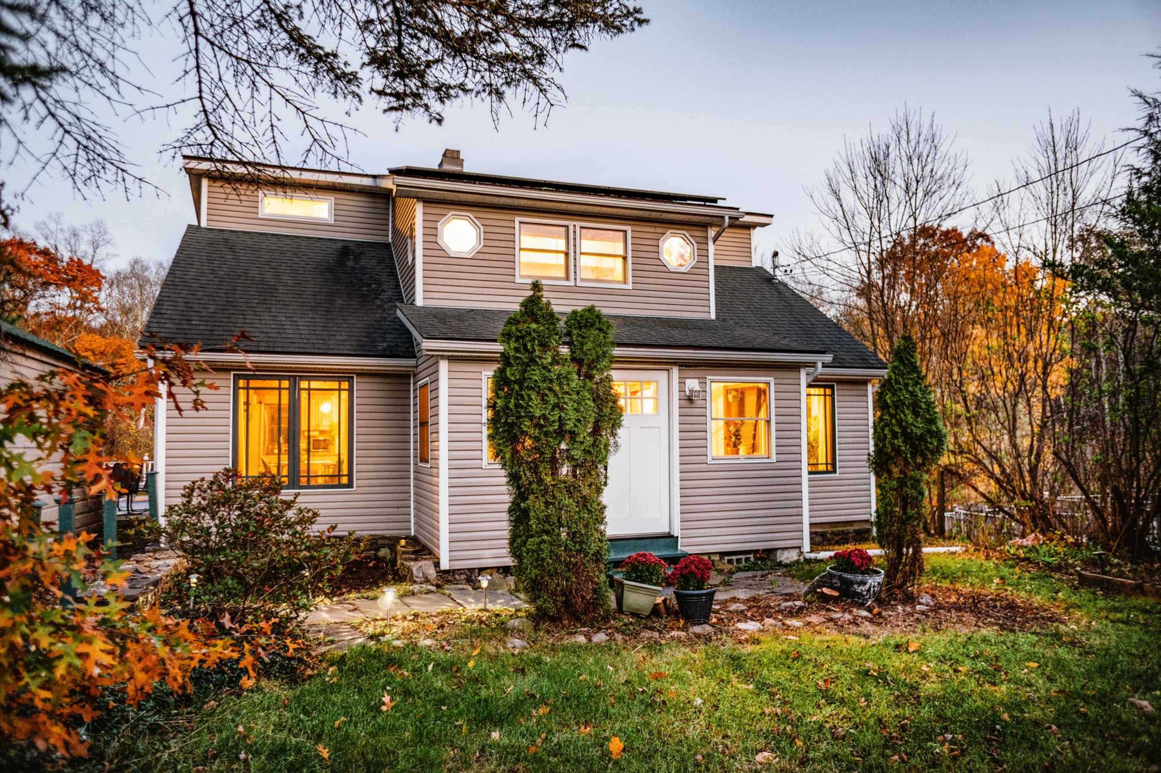 A two-story house with gray siding, surrounded by colorful autumn foliage and illuminated windows at dusk.