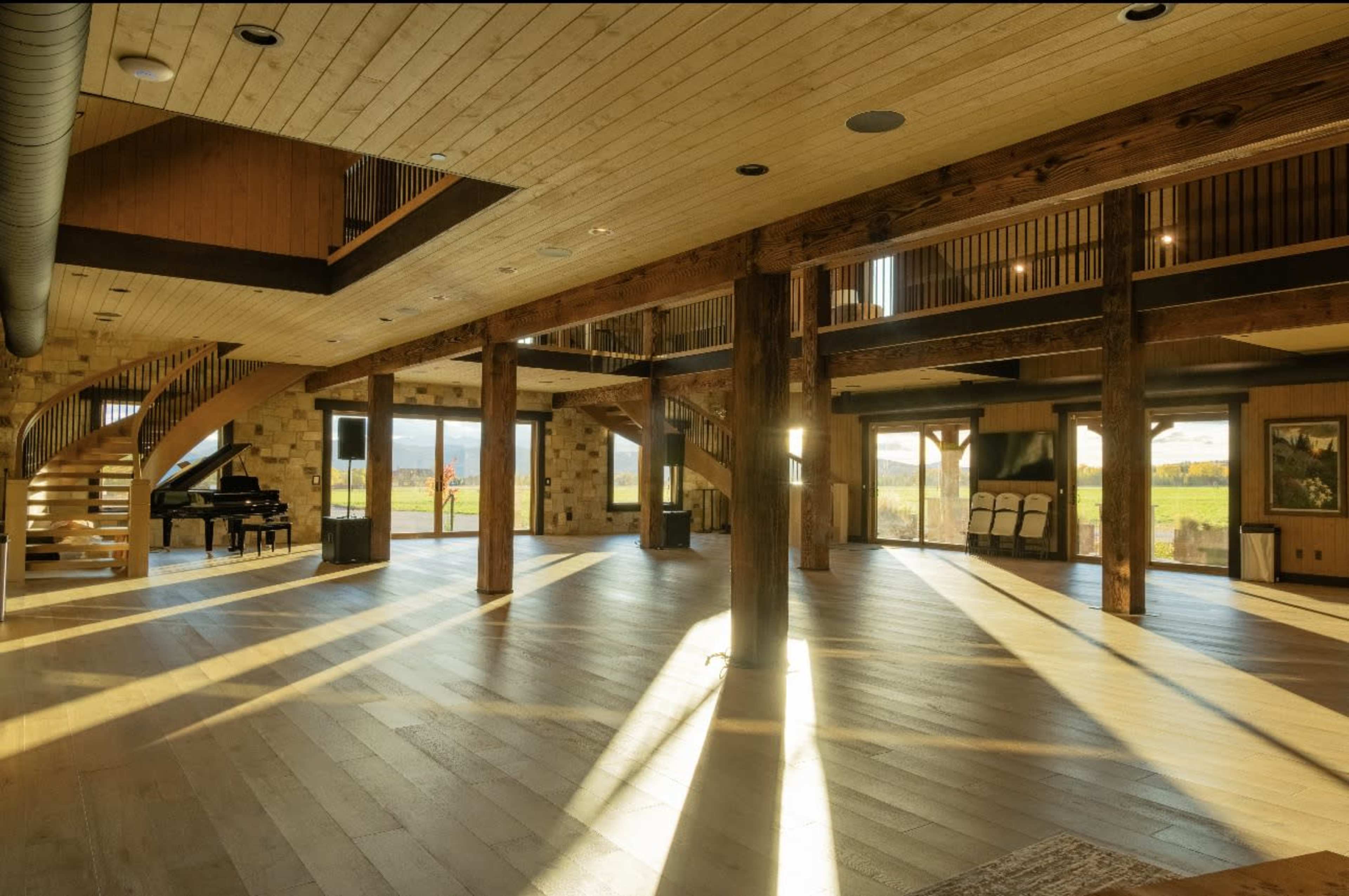The image shows an open, spacious interior of a modern building with wooden beams and a spiral staircase, illuminated by natural light.