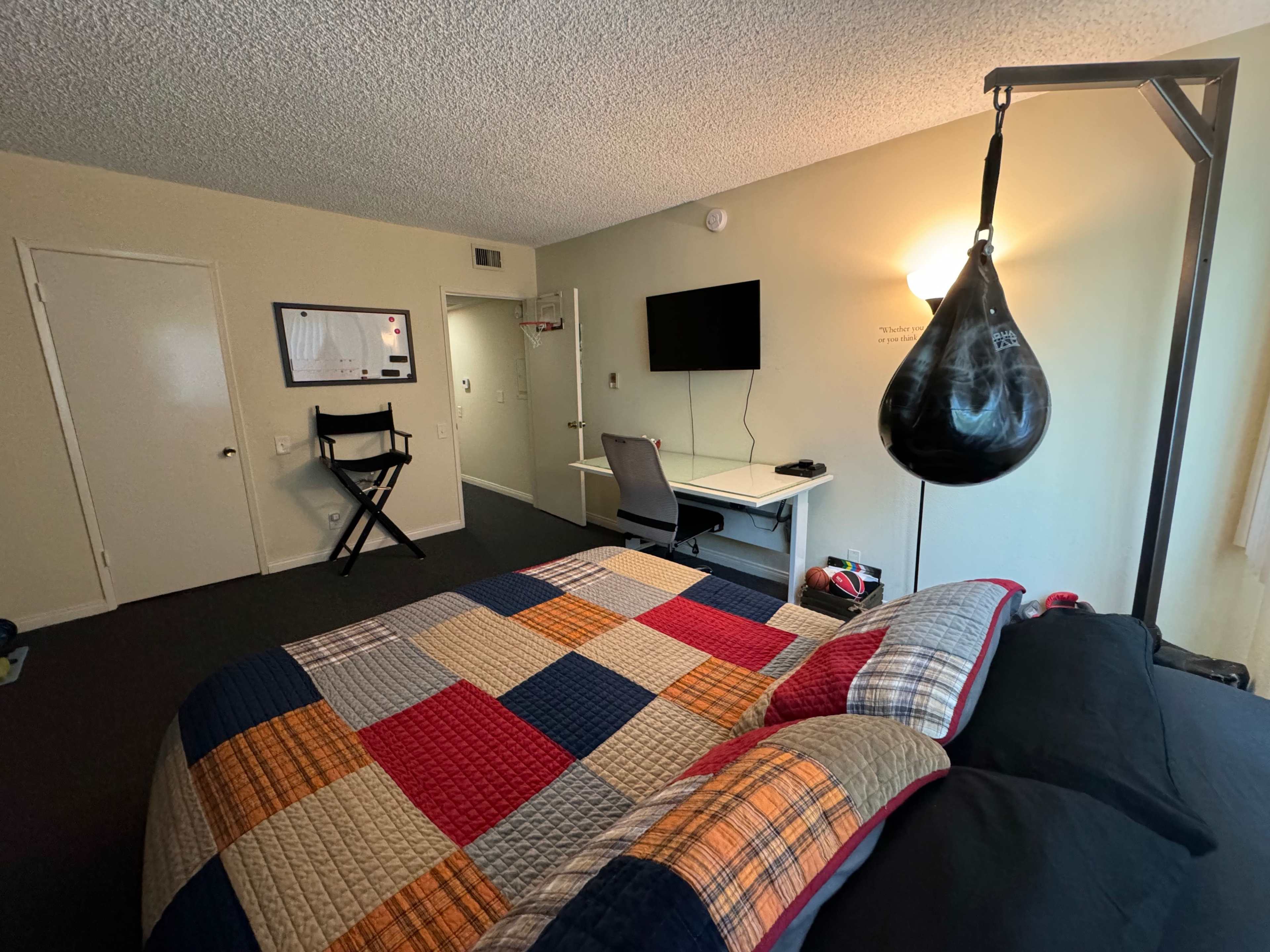 A bedroom with a large bed covered in a patchwork quilt, a desk with a chair and a television mounted on the wall, and a heavy punching bag hanging from a stand.