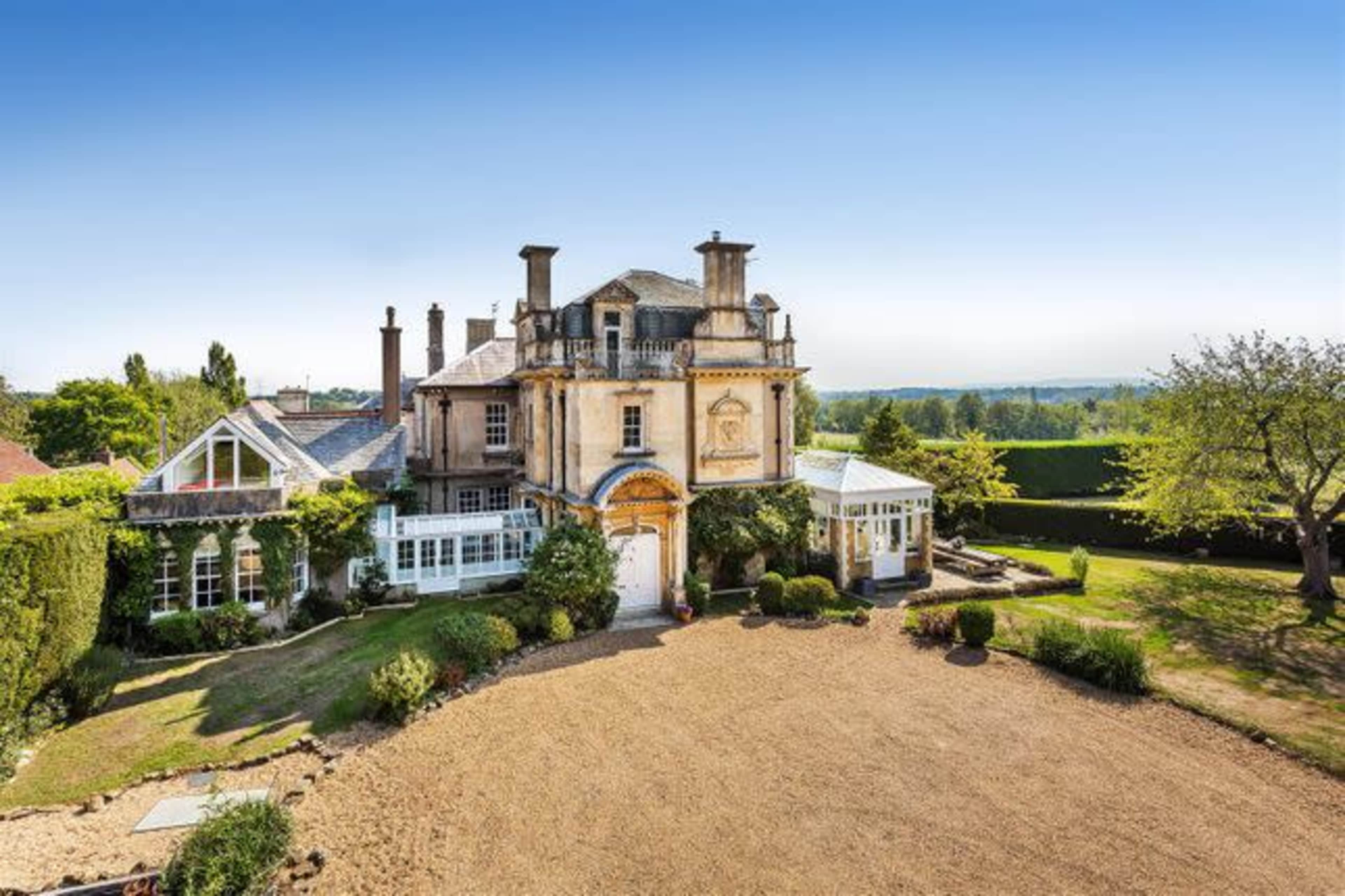 A large, elegant mansion surrounded by well-maintained gardens and trees under a clear blue sky.