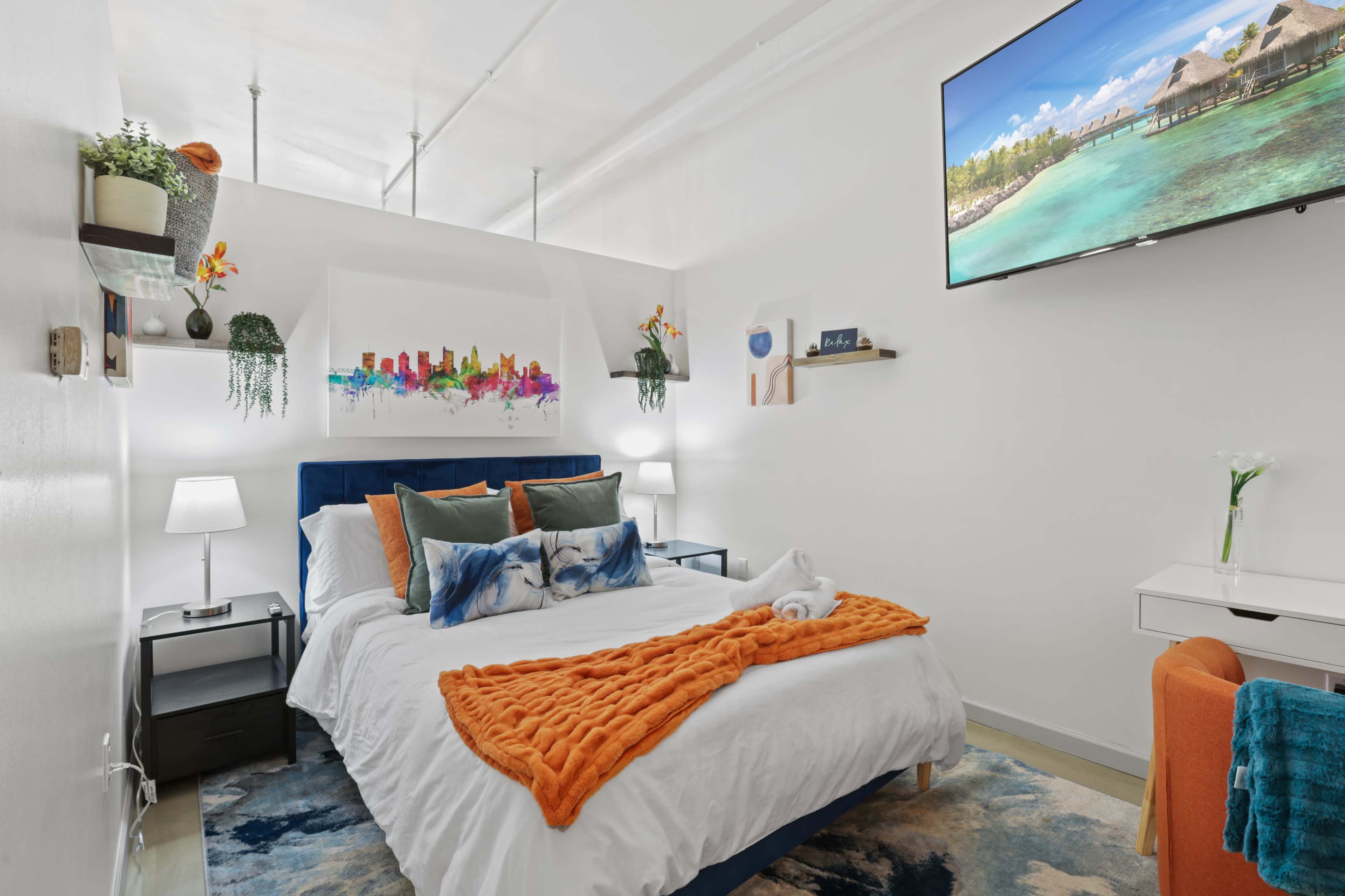 A bright bedroom featuring a double bed with colorful pillows, a television mounted on the wall, and decorative accents including plants and artwork.