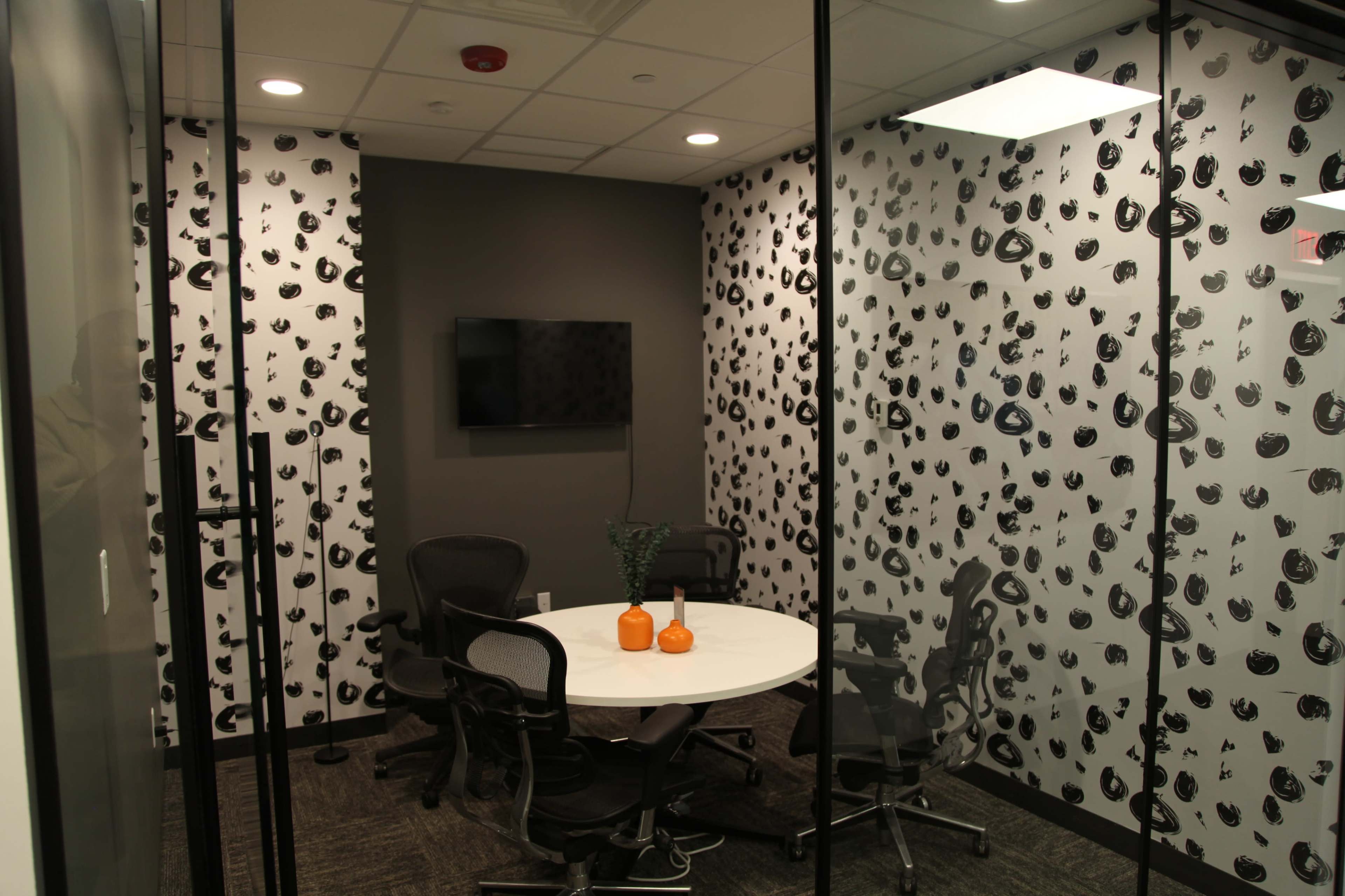 The image shows a glass-walled meeting room with a round white table and four swivel chairs, decorated with black and white patterned walls.