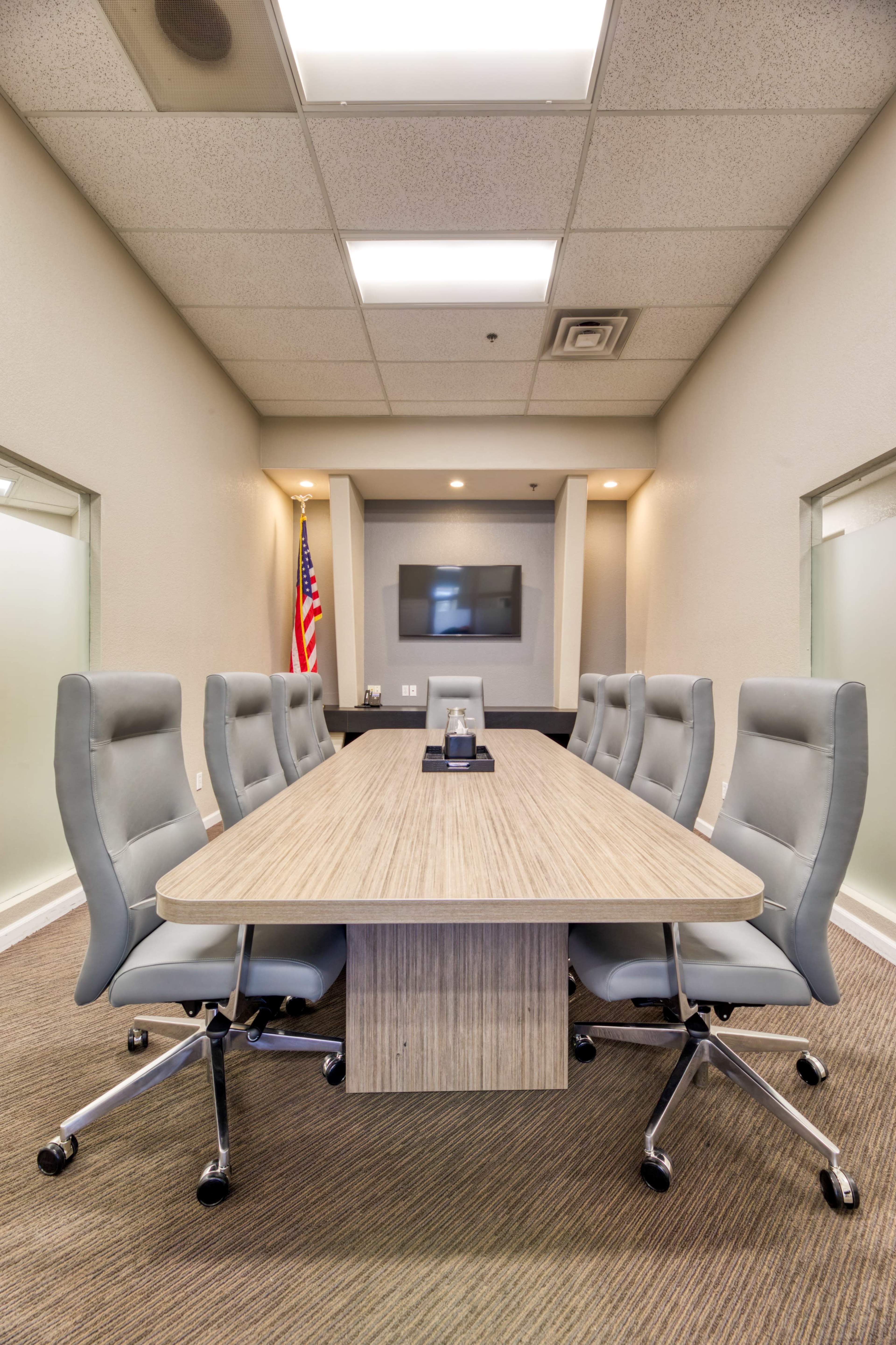 The image shows a modern conference room with a large wooden table surrounded by eight gray swivel chairs, a television mounted on the wall, and an American flag in the background.
