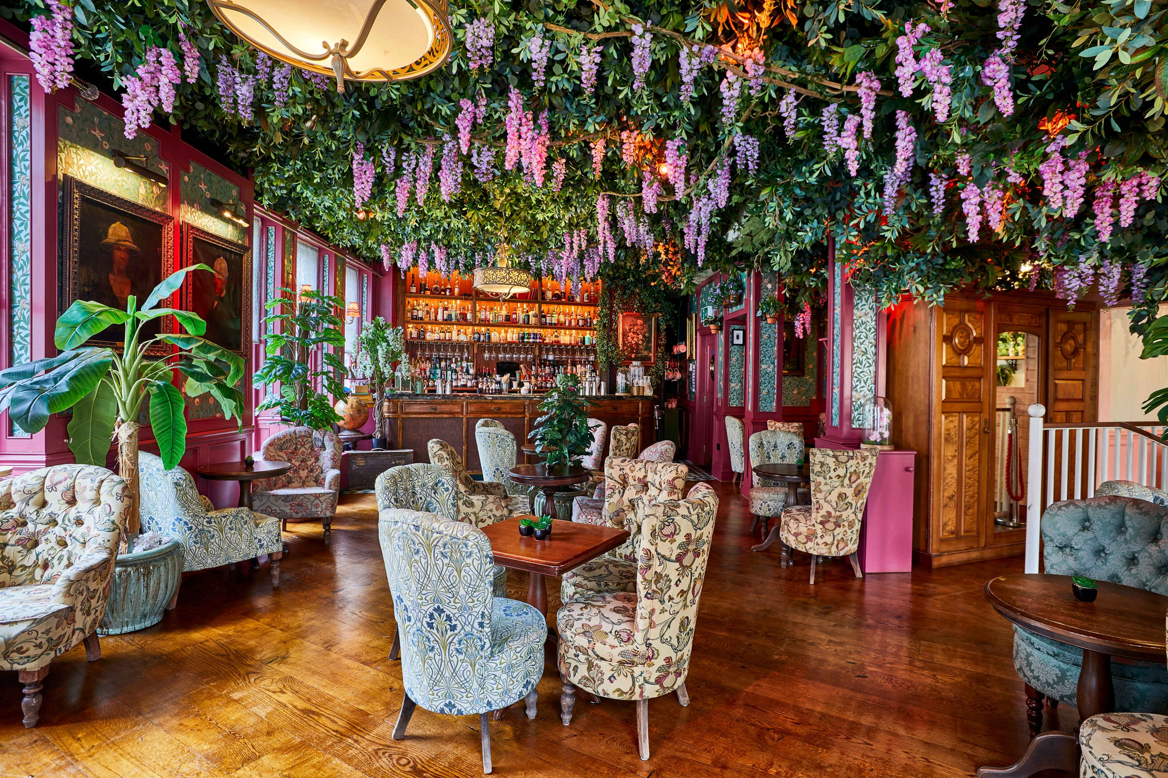 The interior of a vibrant bar features floral decorations hanging from the ceiling, with plush seating arranged around small tables.