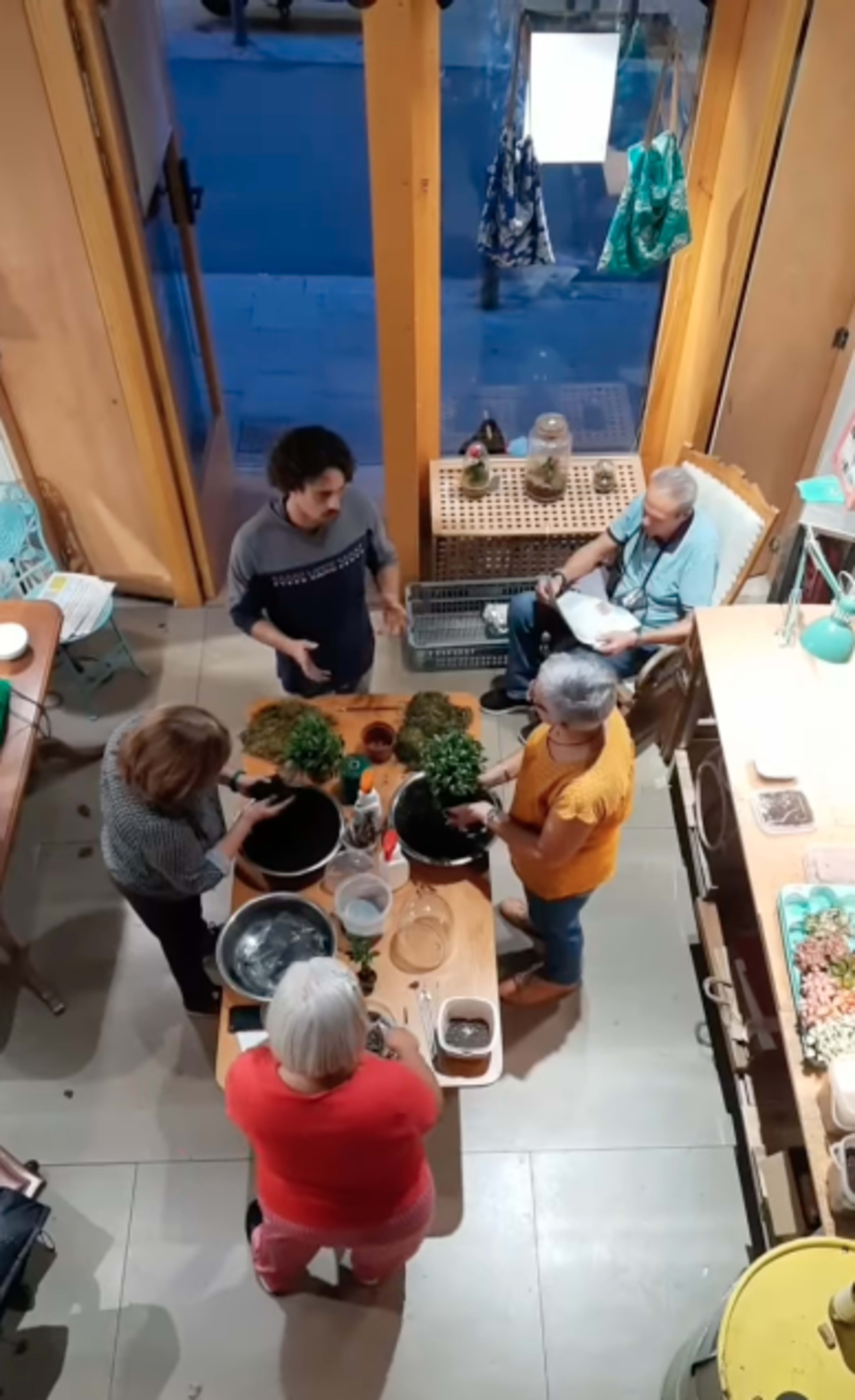 A group of five people is gathered around a table in a brightly lit room, working together on potting plants.
