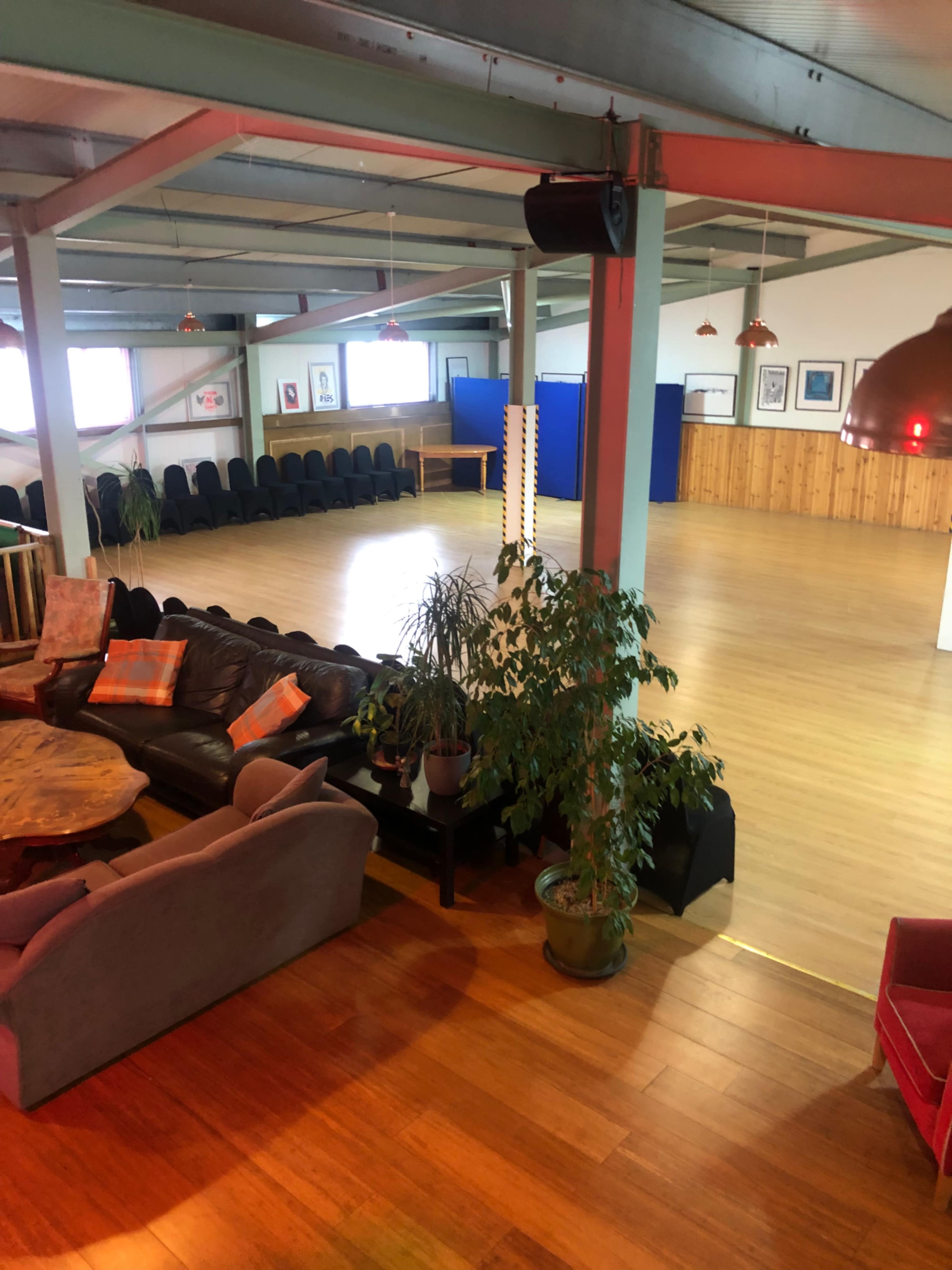 A spacious, empty hall with wooden flooring and seating arranged along one side, accompanied by a few potted plants.