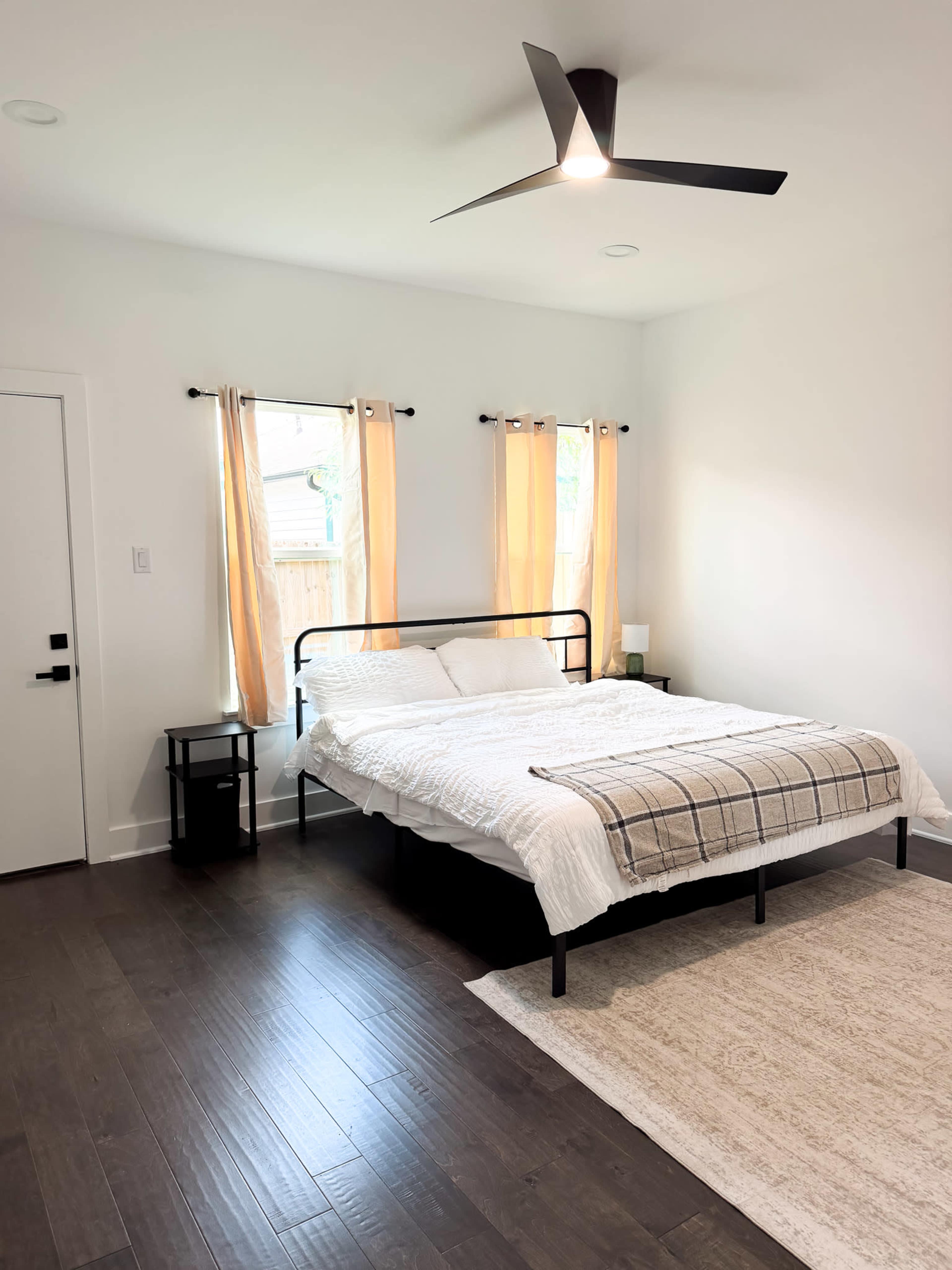 The image shows a simple bedroom featuring a metal bed frame with a white bedspread, two windows with light yellow curtains, a small black bedside table, and a light-colored rug on the wooden floor.
