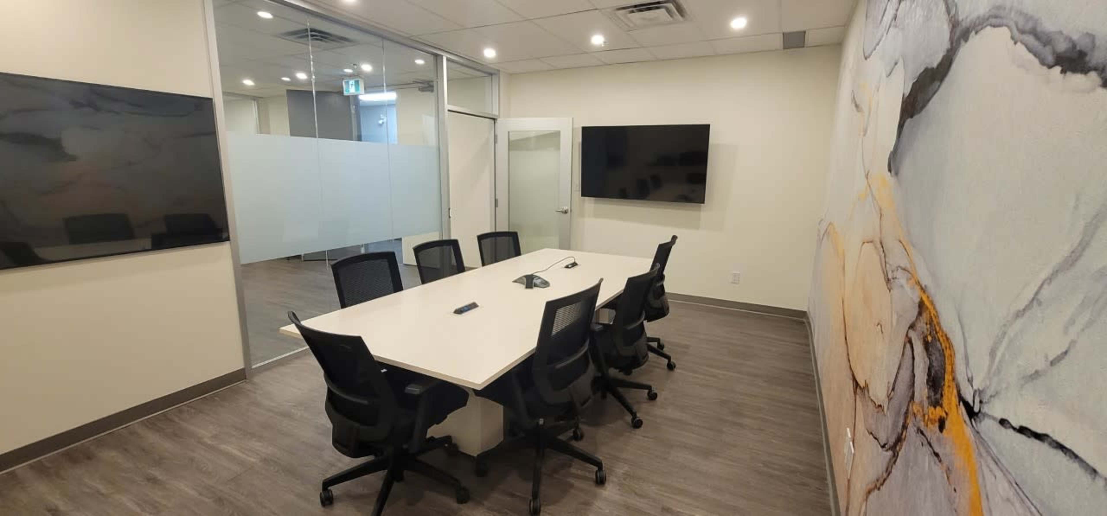 A modern conference room with a long rectangular table surrounded by six black chairs, two wall-mounted screens, and large glass windows.
