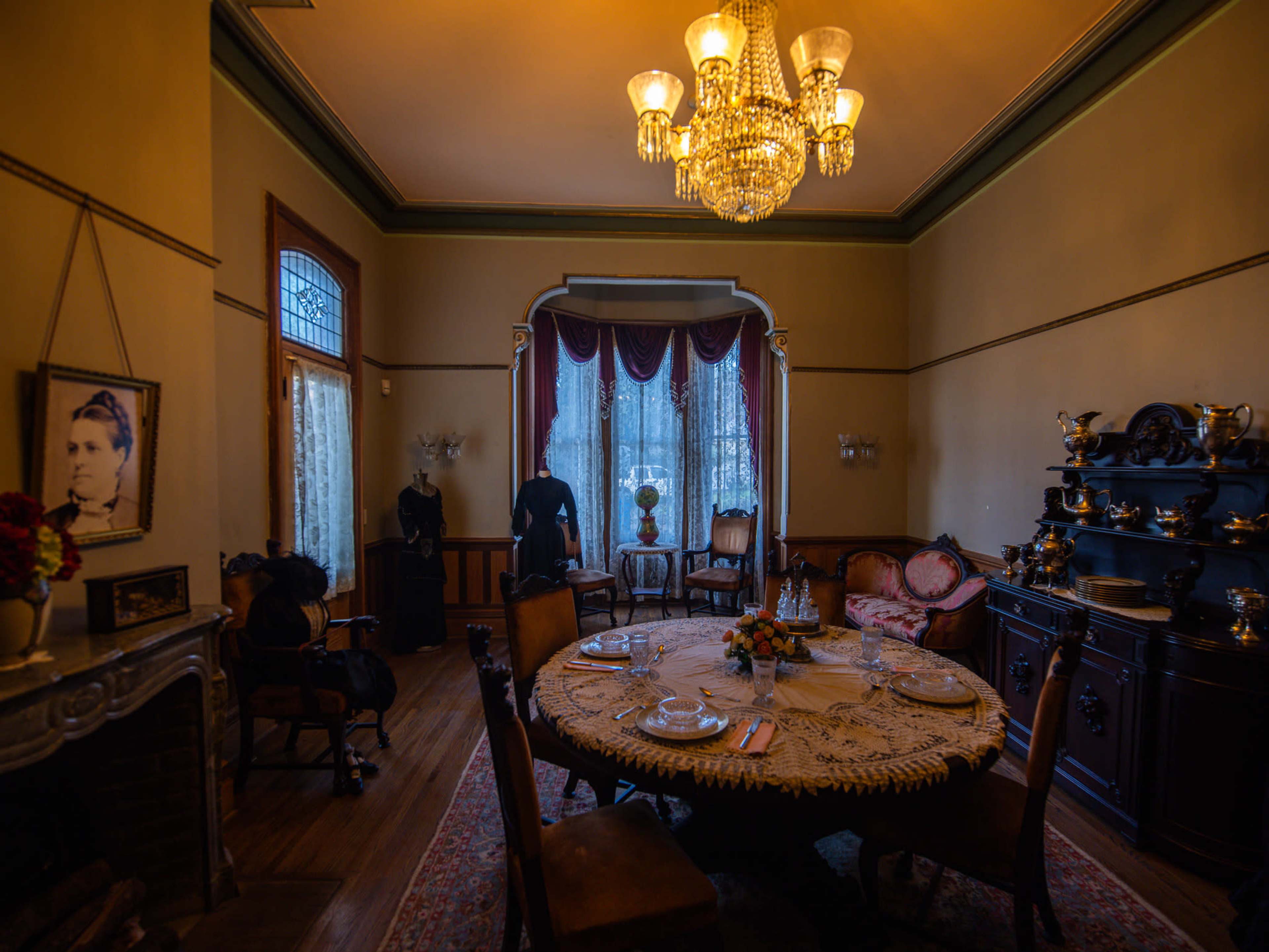 The image shows a vintage dining room with a large table set for a meal, surrounded by ornate furniture and a chandelier.