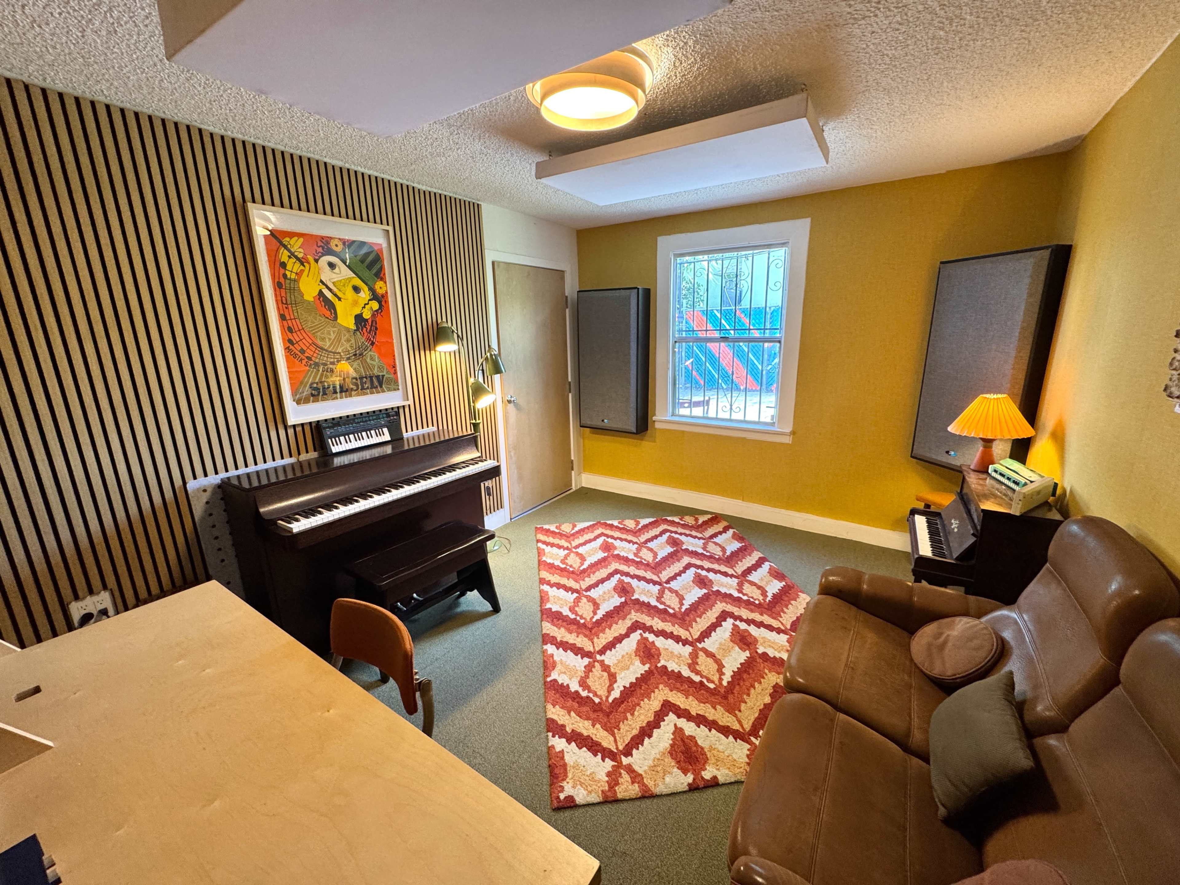 The image shows a music studio with a keyboard, two speakers, a patterned rug, a couch, and yellow walls adorned with wooden paneling.