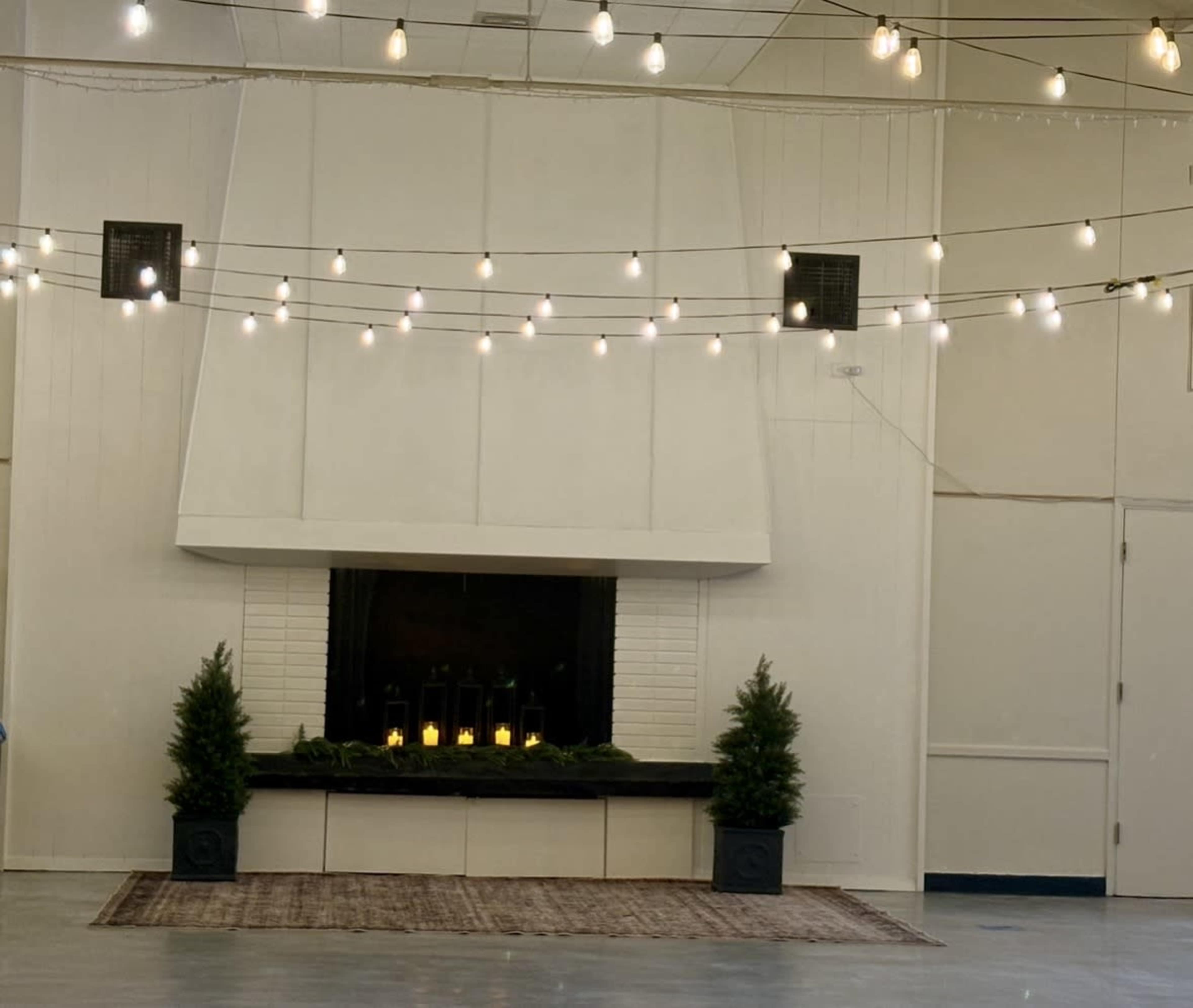 The image shows a spacious indoor venue with a white wall featuring a large fireplace, two potted plants on either side, and string lights arranged above.