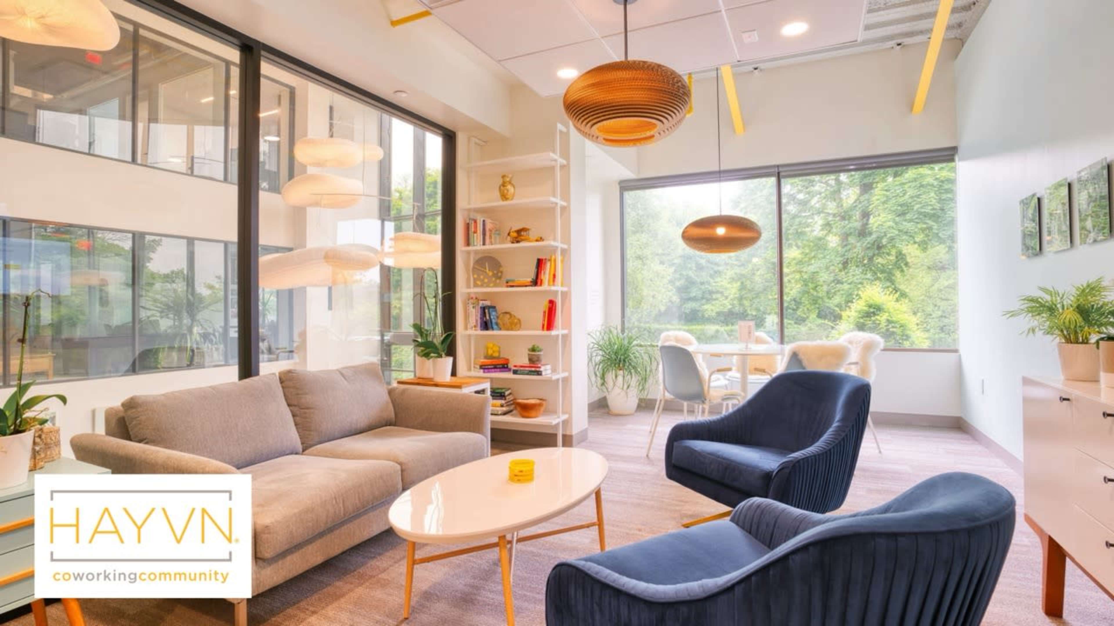 The image shows a modern coworking space with a seating area featuring a gray sofa, two blue chairs, and a round coffee table, along with bookshelves and a dining table visible through large windows.