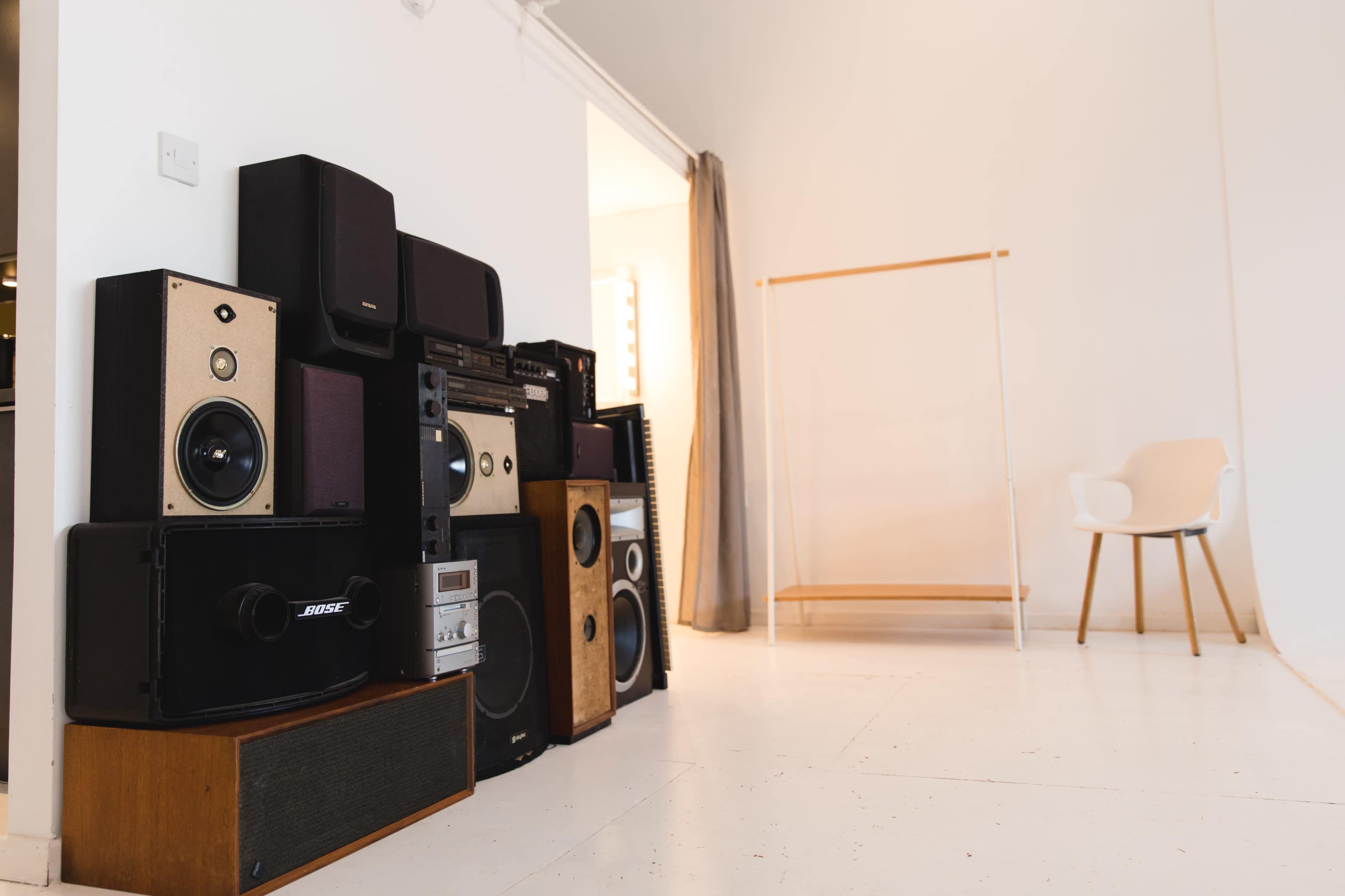 A collection of various audio speakers stacked against a wall in a bright room with a minimalist chair and a clothing rack in the background.