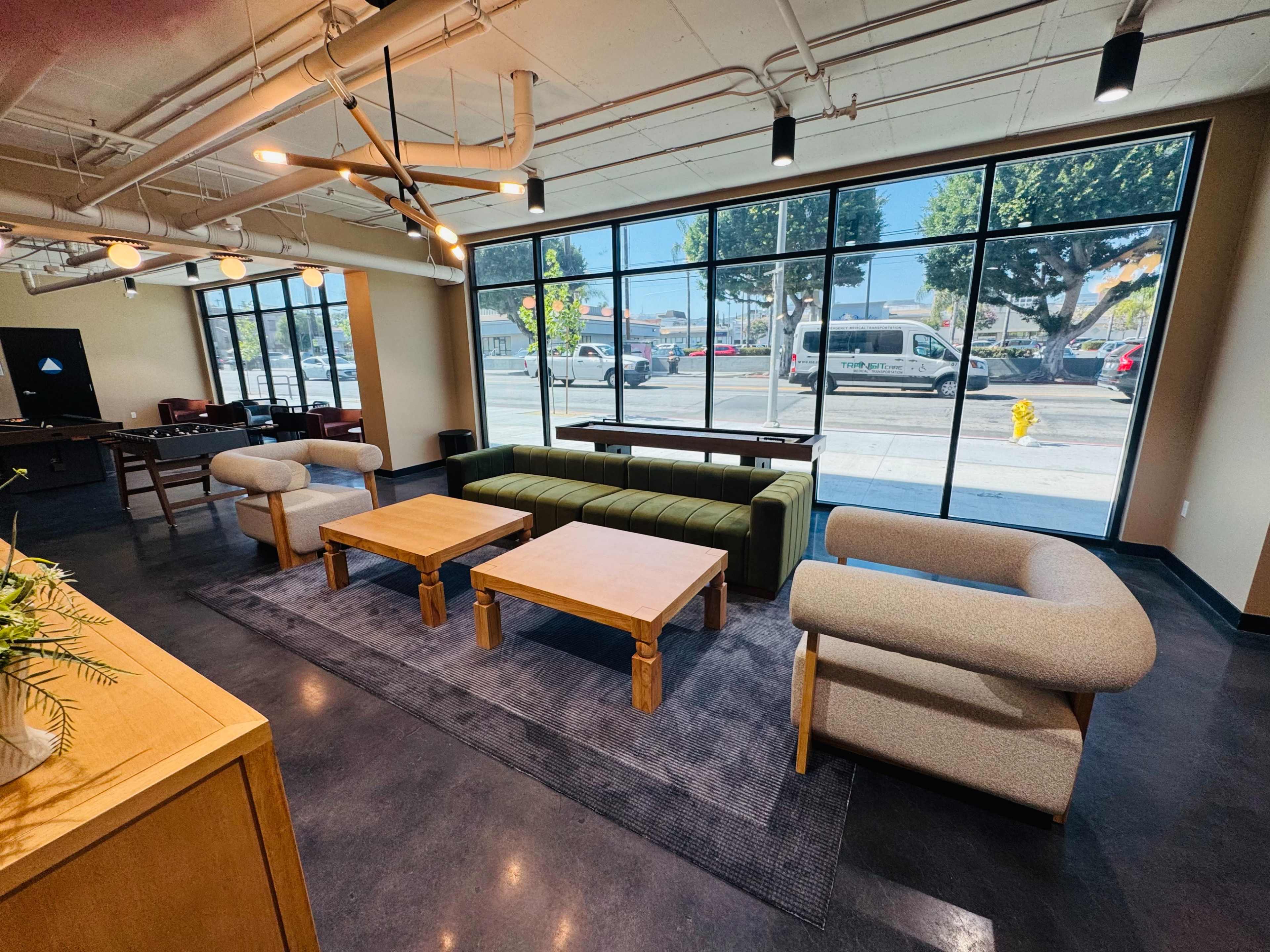 The image shows a modern interior space with a green sofa, wooden tables, and large windows overlooking a street.
