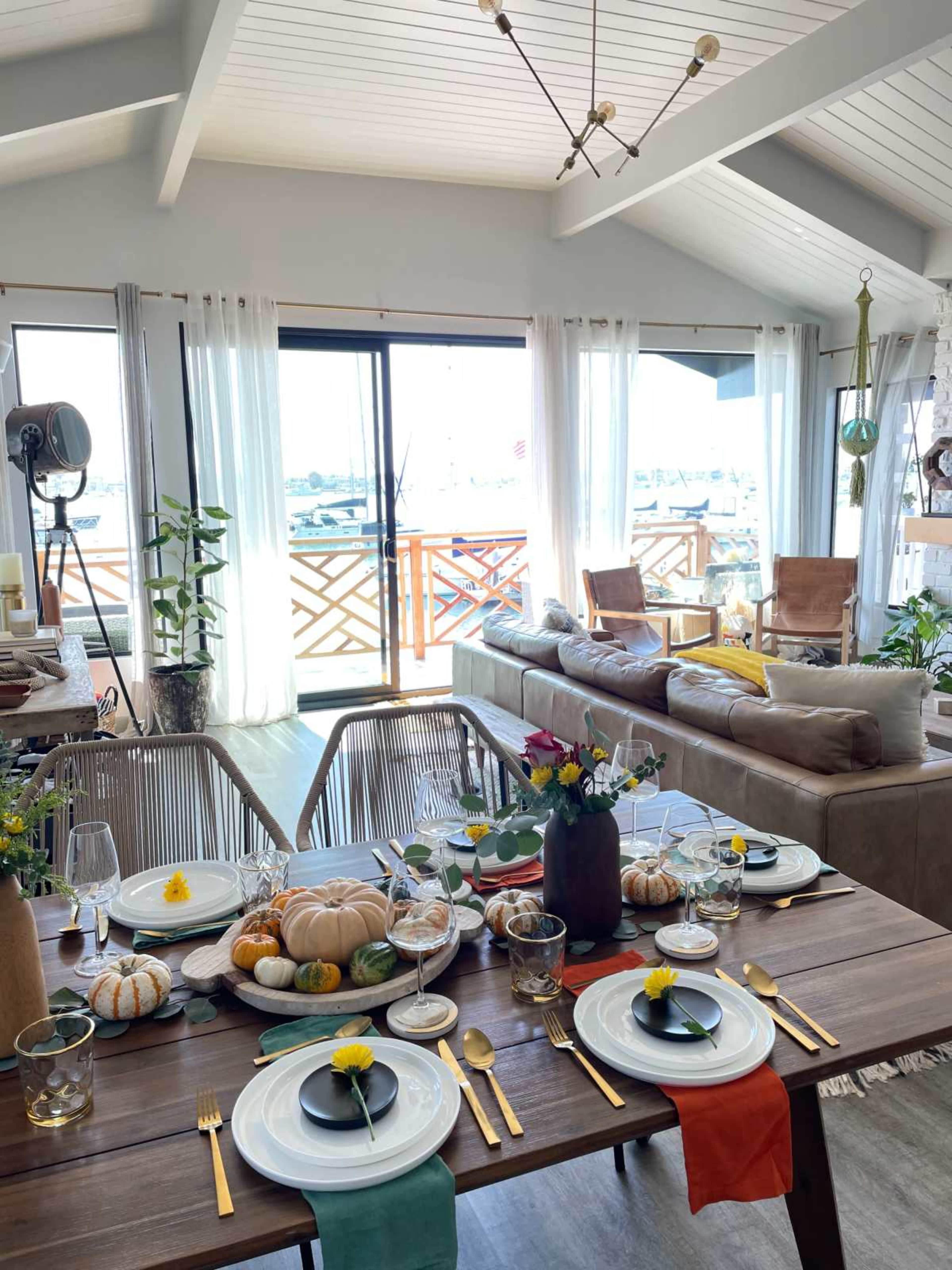 A dining table set for a meal features decorative pumpkins and flowers, surrounded by a bright, airy living space with large windows overlooking a marina.