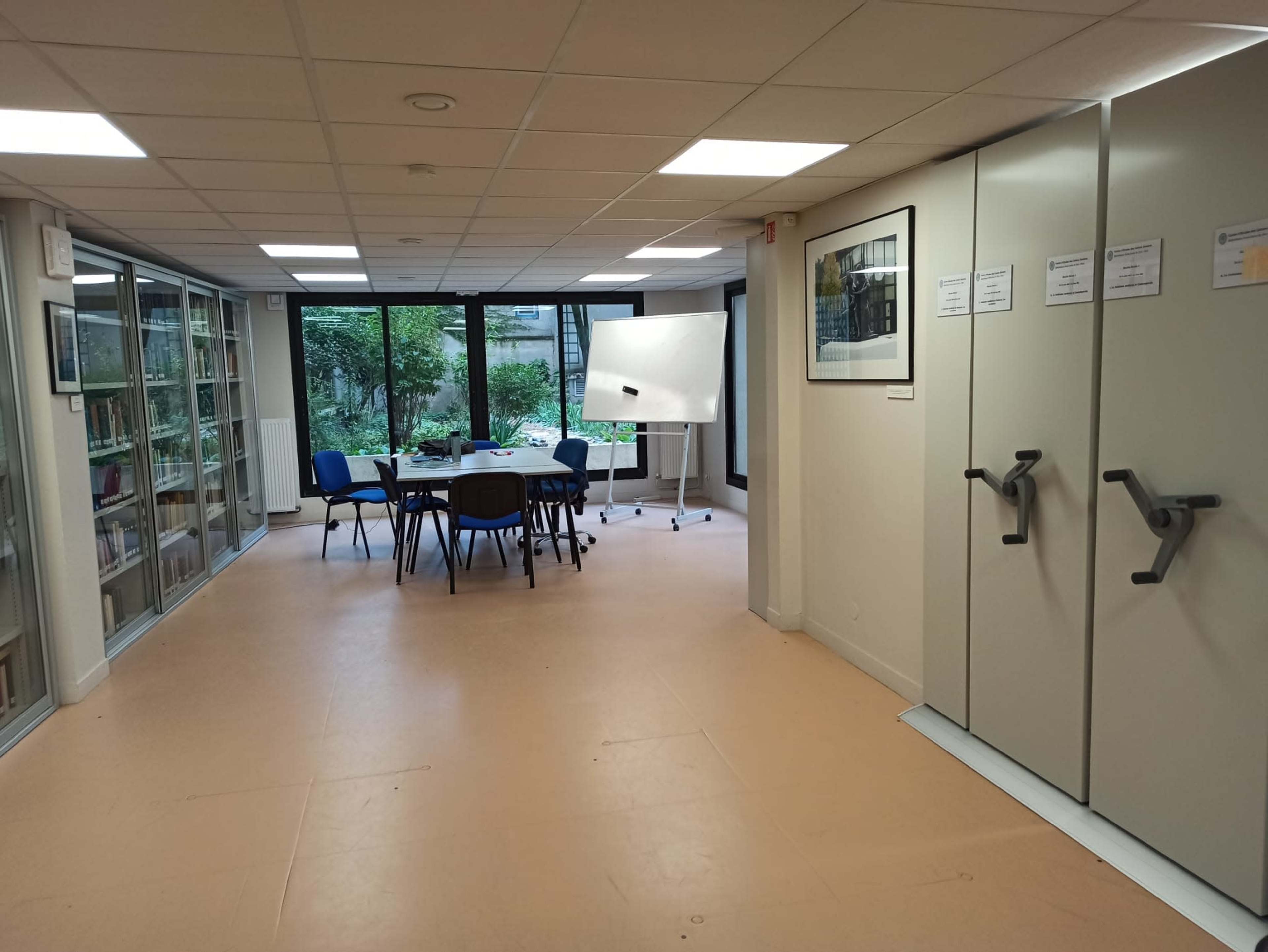 A well-lit meeting room with a round table, blue chairs, a whiteboard, and large windows overlooking a garden.